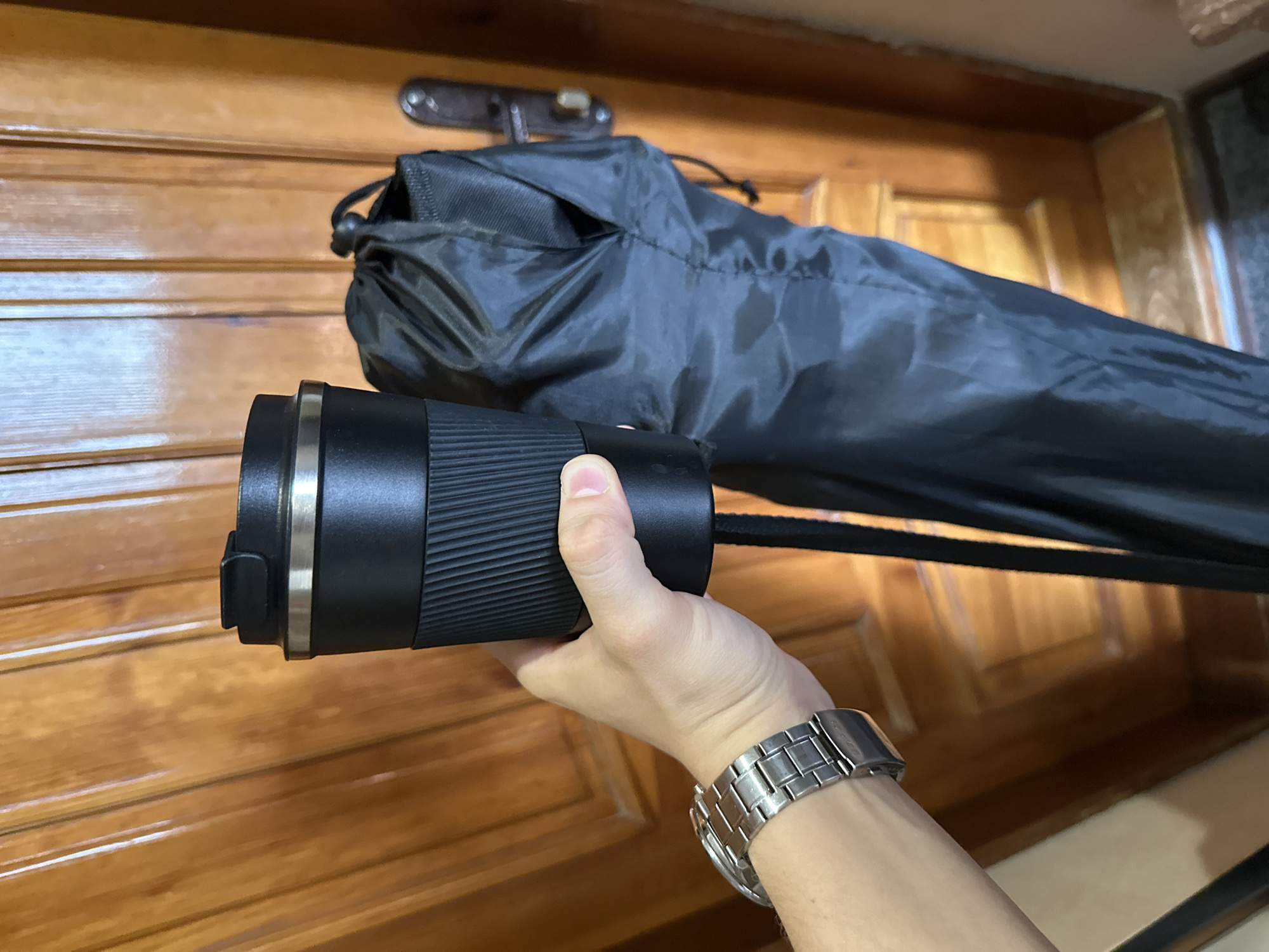 A person wearing a silver watch on their left wrist is holding a black insulated tumbler. Next to the tumbler, and partially obscuring it, is a black drawstring bag. The background is a wooden door with vertical paneling and a dark metal doorknob. The lighting suggests an indoor environment, possibly a hallway or room within a building.