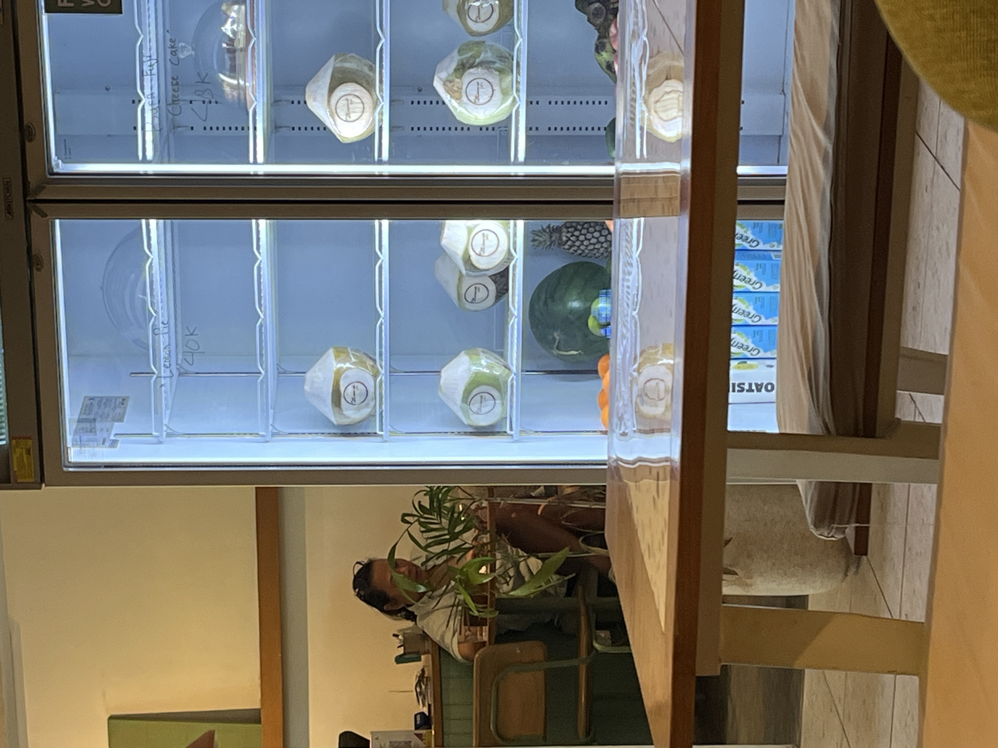 A refrigerated display case, likely within a retail establishment, is prominently featured, containing various beverages and food items including what appears to be coconut water in packaged containers, a pineapple, a watermelon and boxes of "Green" brand drinks. The refrigerated case contains price tags for the items in the refrigerator. A reflection can be seen in the glass, showing a person sitting at a table with a plant nearby. A chair can be seen near the refrigerator as well. The photograph was likely taken in Kuta Selatan, Indonesia.