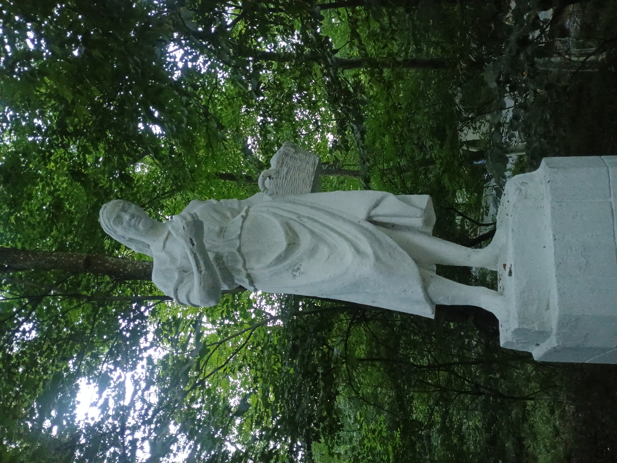 A full-body, low-angle shot captures a white stone statue of a woman from the chest down, standing on a white pedestal. The woman is depicted wearing a long dress and holding a woven basket in her left arm. Her right arm is bent and appears to be holding something in her hand, possibly food or fruit. The statue is set against a backdrop of lush green trees, with sunlight filtering through the dense foliage. The overall impression is of a serene and natural environment, likely a park or a garden, possibly located in the city of Gusar, Azerbaijan, given the context provided. The statue appears to be made of concrete or a similar material, and its white color contrasts with the dark green of the surrounding vegetation. The pedestal is square and has several tiers. The background is dominated by trees, with some dappled light suggesting an overcast or partially shaded day.