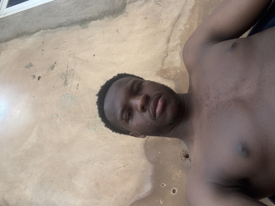 A shirtless, dark-skinned man lies on his back, facing the camera. He has short, dark hair and a slight beard. His eyes are open, and his lips are slightly parted. He is lying on a textured, light brown surface, possibly concrete, with some stains or irregularities. Part of a window is seen in the upper left corner.
