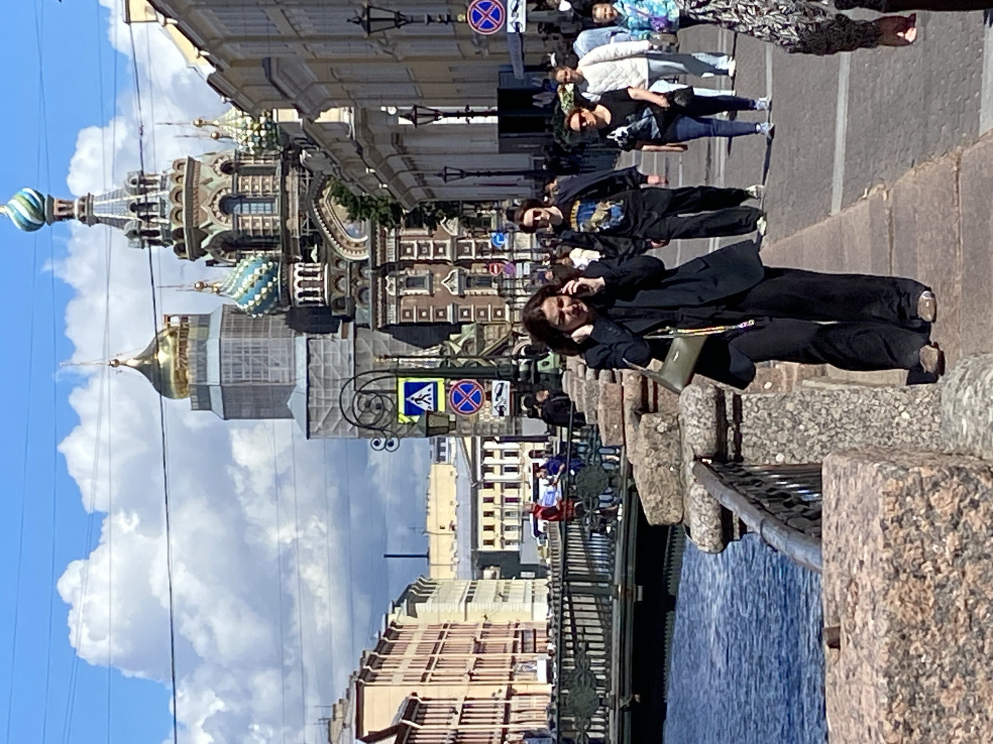 This is a photograph taken on a sunny day in Saint Petersburg, Russia, likely near the Church of the Savior on Spilled Blood. The foreground features a woman in a black suit walking and talking on her phone, with a light green handbag slung over her shoulder. She is on a paved walkway, with a stone wall and body of water to her left. Behind her, other pedestrians are also walking along the same path.
In the background, the distinctive domes of the Church of the Savior on Spilled Blood are visible, some under renovation with scaffolding. The architecture of the church, with its intricate mosaic work and colorful domes, is prominent. Several other buildings, characteristic of Saint Petersburg, line the street. A bridge is visible to the left, spanning the body of water. The sky is blue with fluffy white clouds. Traffic signals and street lamps are also present along the walkway. The overall scene depicts a bustling urban environment with architectural landmarks.