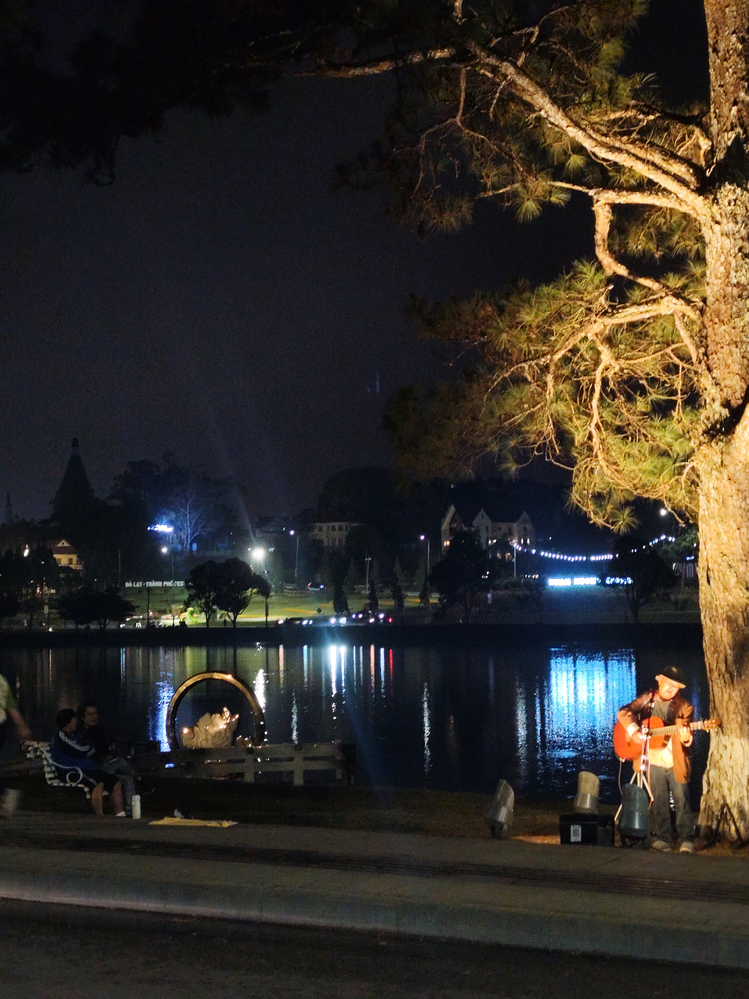 At night in Xuân Hương, Đà Lạt, Vietnam, a man in a hat and jacket plays a guitar next to a tree, illuminated by a spotlight. Two individuals sit on a bench near a circular structure beside a lake reflecting city lights. The cityscape includes buildings, trees, and decorative lighting.