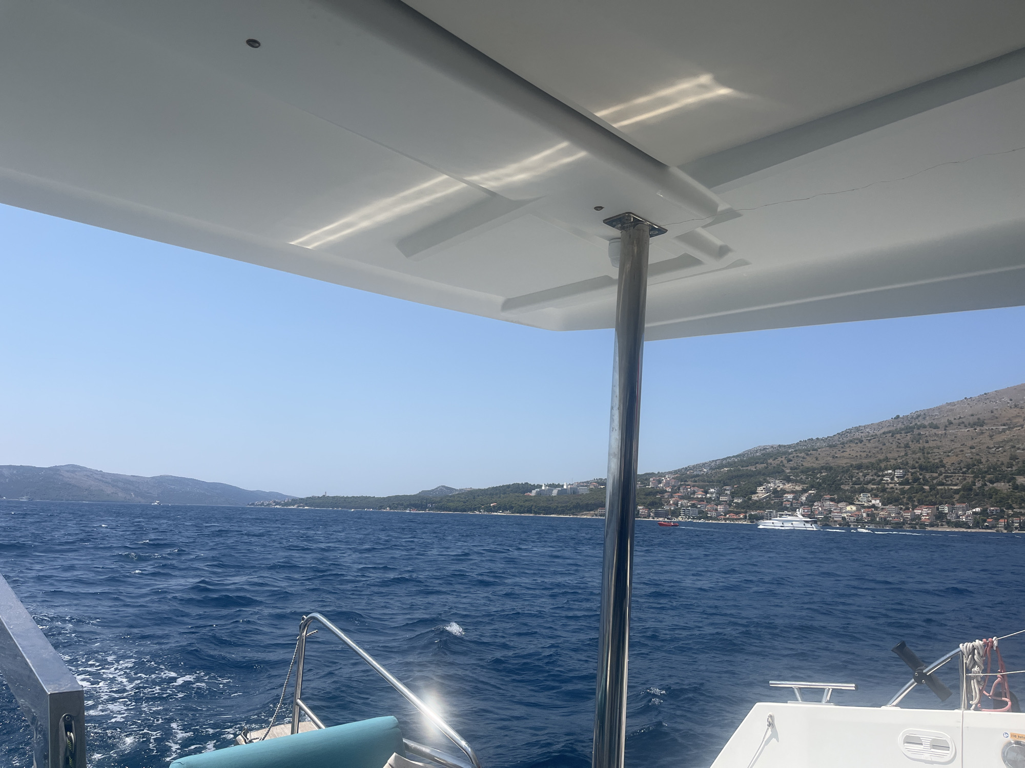 A view from the deck of a boat in the Adriatic Sea, looking towards the coast of Trogir, Croatia. The boat is moving, creating a wake in the choppy blue water. In the foreground, the white canopy of the boat is visible, supported by a metal pole. The coastline in the distance is lined with buildings and dotted with vegetation, with hills rising behind. The sky is clear and blue.