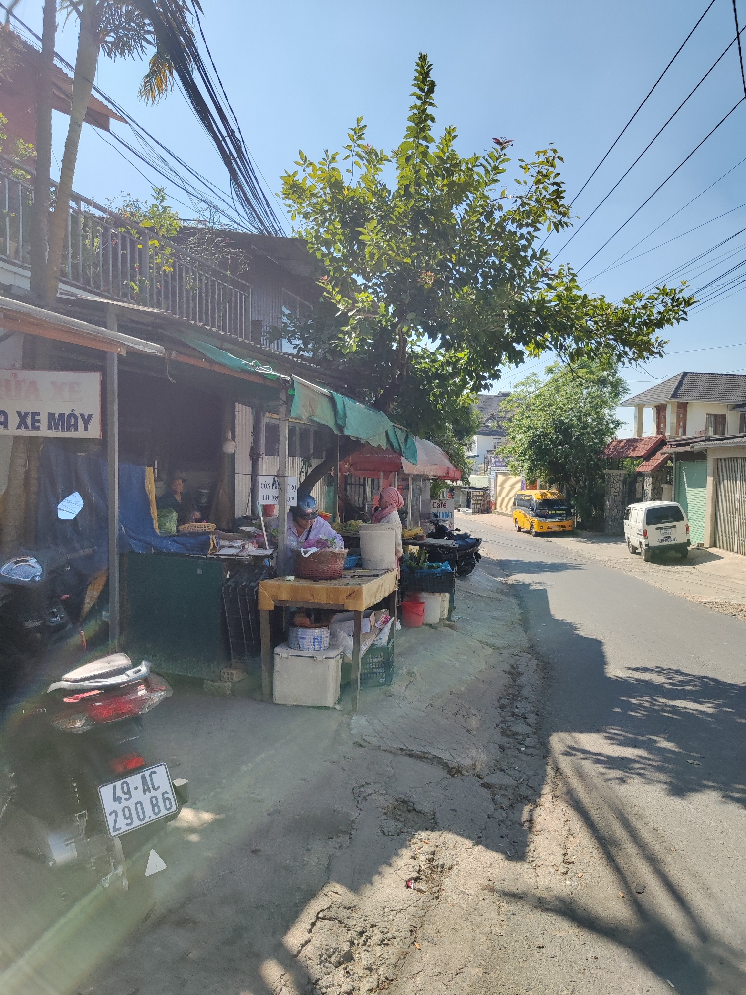 The image captures a street scene in Xuân Hương - Đà Lạt, Vietnam. A small, makeshift shop is set up along the side of the road. A person wearing a helmet is seated inside the shop behind a wooden table. Other shopkeepers are visible in the store. A motorcycle with a license plate reading "49-AC 290.86" is parked in front. The shop's signage, partially obscured, includes phrases like "ỬA XE," "A XE MÁY," "CÒN CHA TRO" and "LH: 035 379". Another signage partially visible is "Café". A yellow vehicle is parked further down the street, along with a white van. The road is paved and shows signs of wear and tear. Trees line the street, casting shadows on the road. Overhead power lines are visible. Buildings are visible in the background.