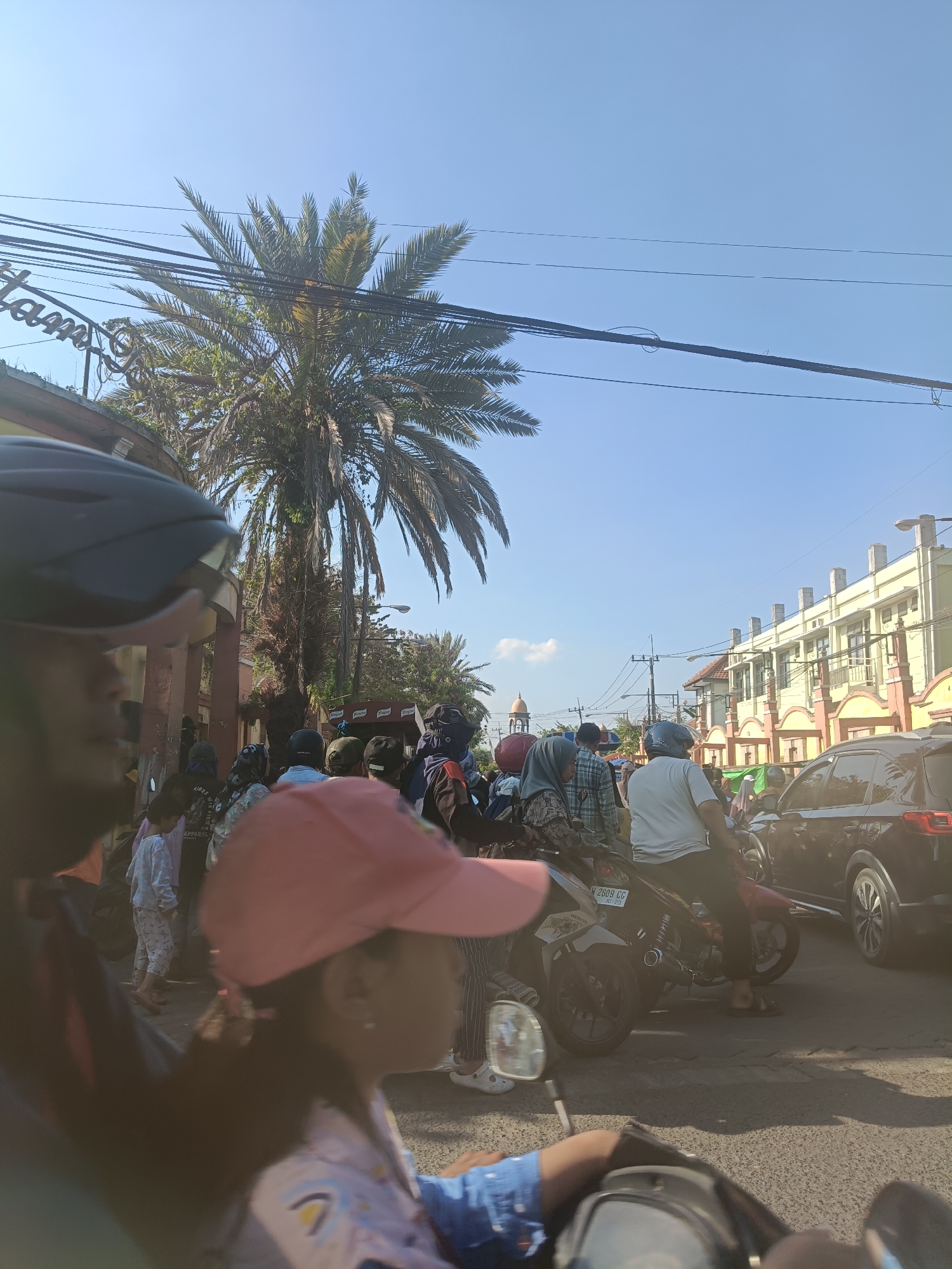 The image captures a street scene in Kembangan, Indonesia, under a clear blue sky. A tall palm tree dominates the left side of the frame, its fronds reaching towards overhead power lines. In the foreground, a person wearing a pink hat is partially visible, suggesting a street-level perspective. Numerous individuals, many on motorcycles, are gathered on the street, indicating potential traffic or a local event. Some are wearing helmets and casual clothing, with a mix of genders and ages present. A black car is visible on the right side of the road, partially obscuring other vehicles. In the background, a building with a light yellow facade and red accents stands prominently, and a golden dome structure, possibly a mosque, is visible in the distance. The overall impression is of a busy urban environment on a sunny day.