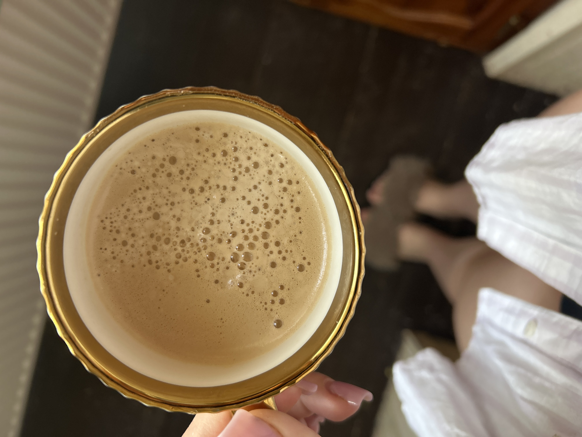 This image shows a top-down view of a delicate porcelain teacup with a gold rim, filled with a frothy beverage, likely coffee or tea. A person's hand, with neatly manicured nails, is holding the cup from the left side. In the background, out of focus, are a person's legs clad in fluffy slippers, and a white garment, possibly a shirt or dress. The setting appears to be indoors, with dark wooden flooring visible. The image does not contain information that would allow for a definitive identification of its location within the city of Basarabeasca, Moldova.