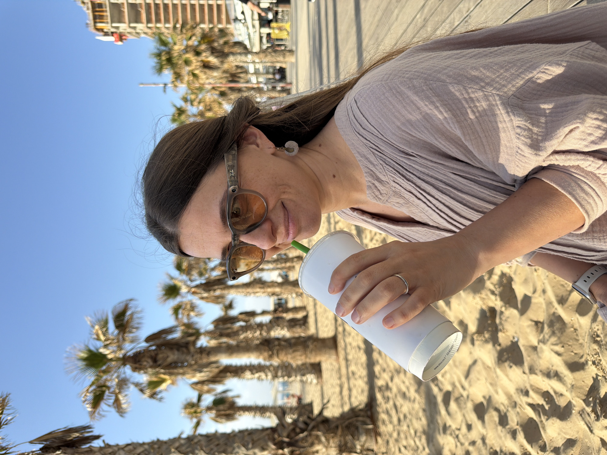A woman in sunglasses drinks from a white cup with a green straw on a sunny day in Bat Yam, Israel. She wears a light purple shirt, an earring on her left ear, and a ring on her left ring finger. A white smartwatch is visible on her left wrist. Palm trees are in the background. The ground is sandy, suggesting a beach location. Buildings are visible in the background. The photo appears to have been taken from a low angle.