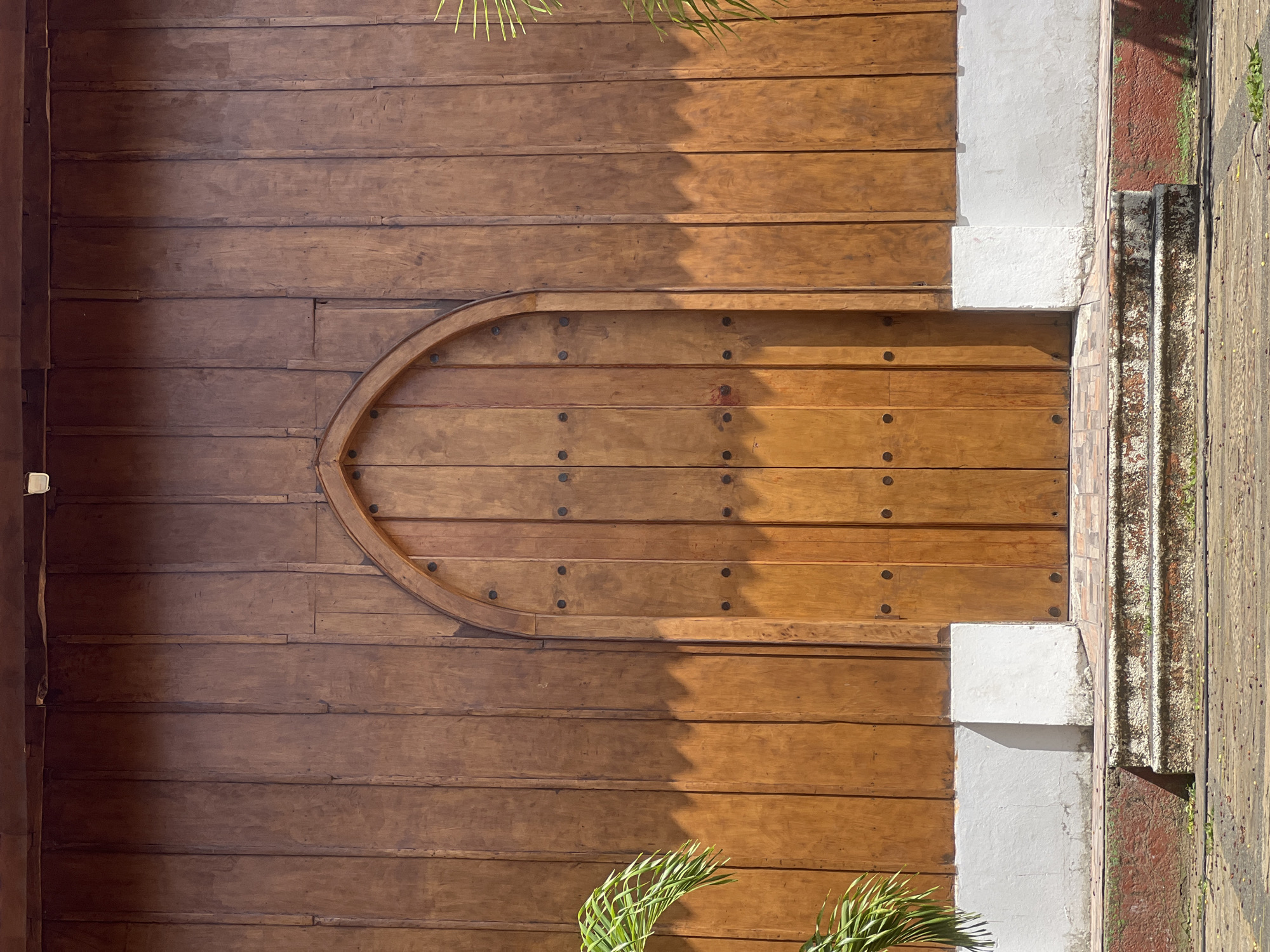 This is a photograph of a large, arched wooden door set into a wooden wall. The door is made of vertical planks and has decorative dark metal studs. Above the door, the wooden wall is shaded, casting a distinct shadow pattern. The bottom of the wall has a white base. In front of the door are several stone steps leading up from a paved area. To the left and right of the door, palm fronds are visible, suggesting an outdoor setting. The overall impression is one of an entrance to a building with a rustic or traditional architectural style, likely in a warm climate given the foliage. This scene is located on the exterior of the San Juan del Sur Cathedral in San Juan del Sur, Nicaragua.
