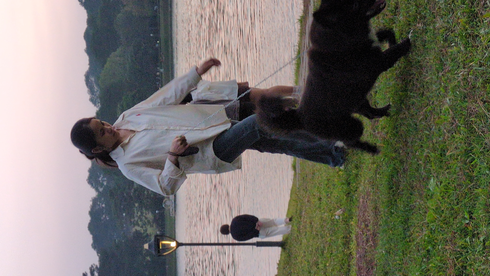 The image captures a scene at dusk near Xuan Huong Lake in Da Lat, Vietnam. A woman in a light-colored long-sleeve shirt and jeans holds a dog leash. She appears to be struggling with a black dog that is pulling on the leash and jumping on a grassy area next to the lake. In the background, another person stands next to a lamppost along the lake's edge. Trees line the horizon across the lake. The water surface reflects the fading light of the sky.