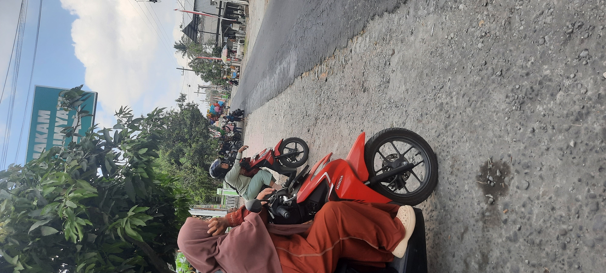 In Jatitengah, Indonesia, two women are seen sitting on motorbikes on the side of a rural road. The woman on the left, wearing a pink hijab and an orange outfit, is looking away from the camera, her hand resting on her chin. The woman on the right, wearing a blue helmet and a light blue outfit, is looking forward. Both are on red Honda motorbikes. The road is paved but has a rough texture and is lined with gravel and some puddles. Trees and greenery are visible on the left side of the road, and a sign reading "MAKAM IMY..." is partially obscured by the foliage. On the right side of the road, there are buildings, a few stalls, and more motorbikes. The sky is partly cloudy.
