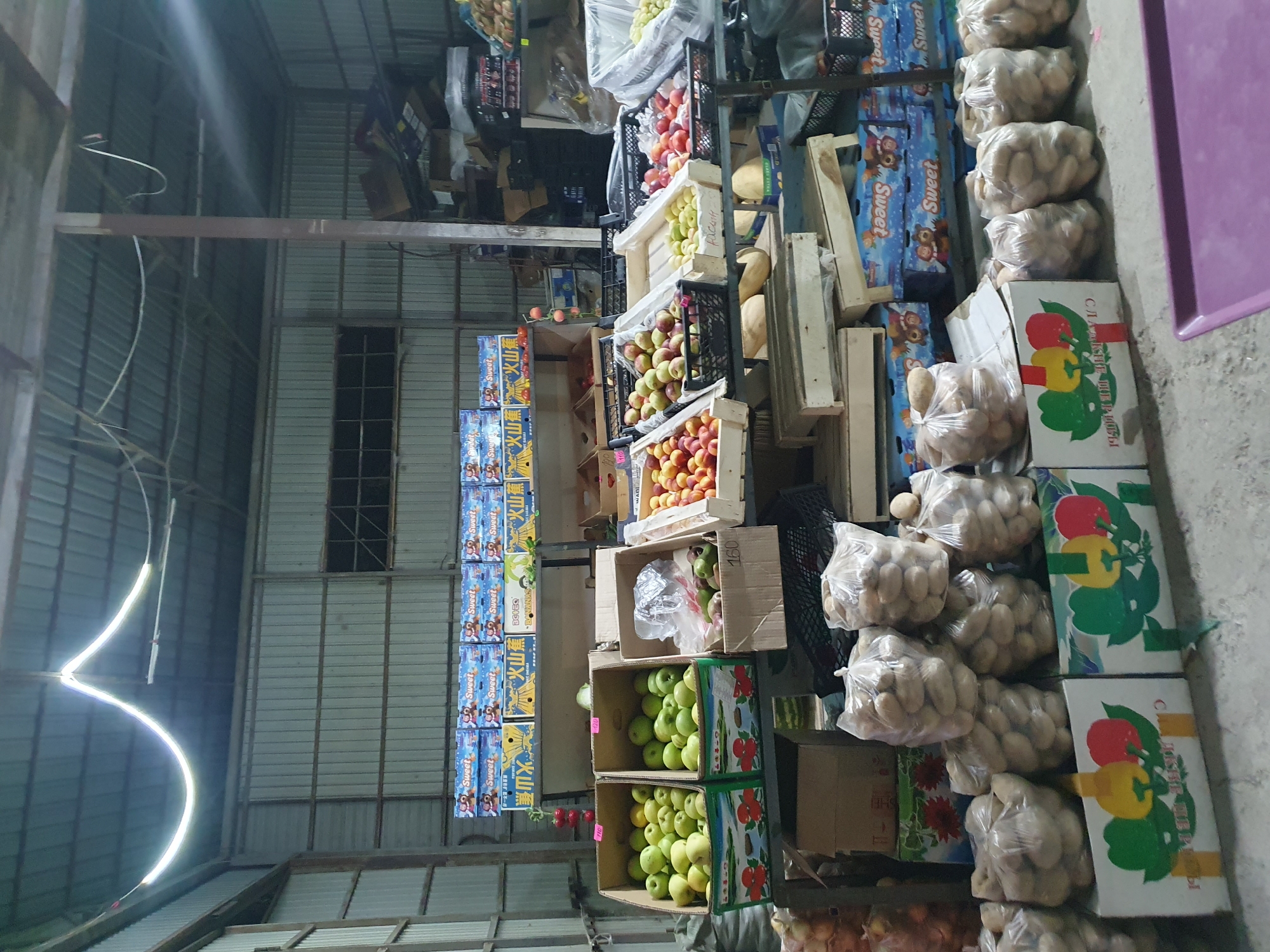 The image depicts the interior of a wholesale produce market, likely located in Maevka, Kyrgyzstan, judging by the Cyrillic script visible on some packaging. The scene is dimly lit, with a prominent fluorescent light fixture hanging from the corrugated metal ceiling. The environment is utilitarian, with exposed metal walls and a concrete floor.
The central focus is a display of fresh produce. On shelves and stacked on the floor, various fruits and vegetables are organized for sale. Boxes of apples, peaches, and possibly other fruits are visible, along with bags of potatoes. Some of the produce is in cardboard boxes decorated with colorful images of vegetables, and other items are in plastic bags. There are also stacks of cardboard boxes, some labeled with the word "Sweet" and featuring cartoon bear imagery, suggesting they are packaging for a product.
In the background, more boxes and crates are stacked, indicating a storage or distribution area. The overall impression is one of a working market, with goods neatly arranged for commercial transaction. No people are visible in the image.