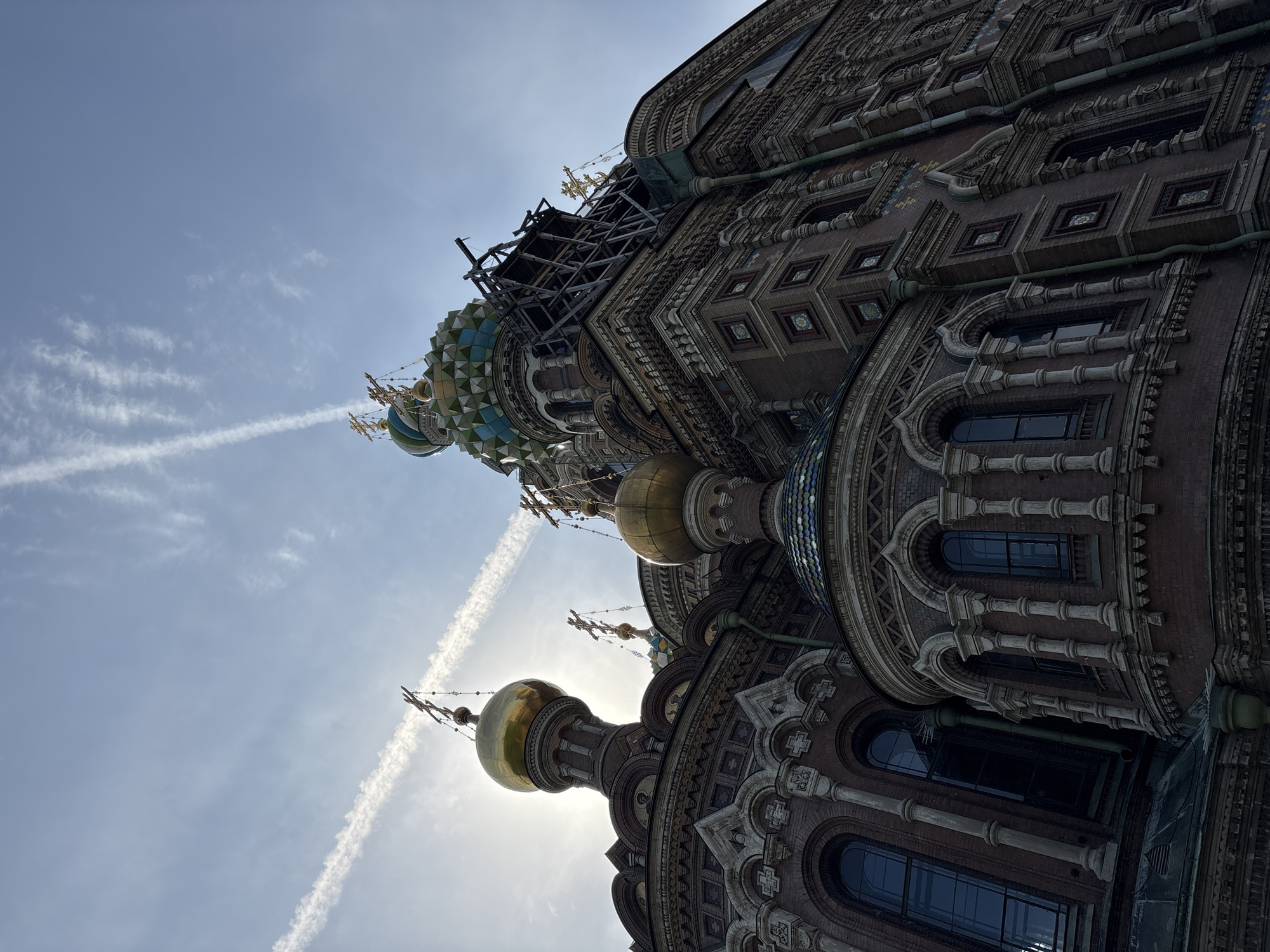 The image depicts the Church of the Savior on Spilled Blood in Saint Petersburg, Russia, from a low-angle perspective. The architecture features multiple domes, including a gold one silhouetted against the sun. The sky above is pale blue with contrails. Scaffolding is visible near the top of one of the domes. The church is richly ornamented with brickwork, arches, and decorative elements. Windows are present.