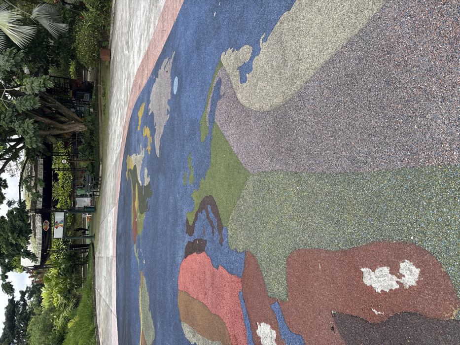 The image displays an outdoor recreational area, specifically the "Mapa Mundial" section within Bioparque Ukumarí, Pereira, Risaralda, AMCO, Colombia. The foreground is dominated by a large, circular, multi-colored map of the world rendered on a rubberized ground surface. Continents are depicted in shades of red, green, brown, light blue, grey, and yellow, contrasting with dark blue sections representing oceans. Visible wear is present on several areas of the map.
A light-colored concrete pathway separates the map from the midground. In the midground, a partially visible covered structure with a dark, possibly thatched roof functions as an entrance or kiosk. A sign featuring a stylized, multicolored bird graphic is mounted on this structure. One individual, attired in dark clothing, is observed standing near the entrance. A dark-colored bench is positioned on the concrete path to the right of the entrance. The background consists of dense tropical vegetation and large trees under an overcast sky.