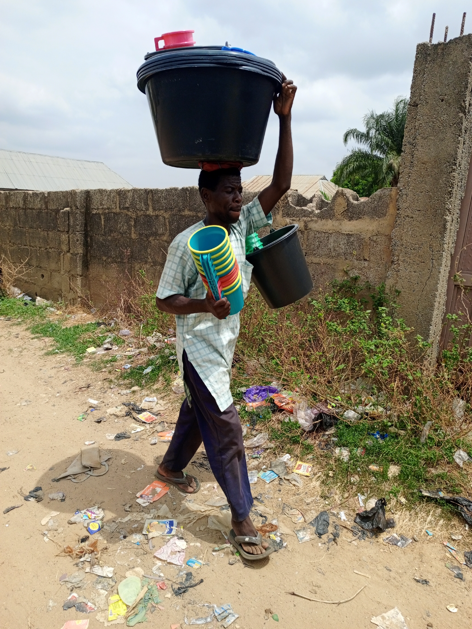 In Keffi, Nigeria, a man carries a stack of colorful plastic cups in his left hand and a black bucket in his right. He balances a larger black bucket on his head. The man wears a light-colored, checkered, short-sleeved shirt, dark trousers, and sandals. He walks on a dirt path strewn with litter. Behind him is a concrete block wall with vegetation and refuse growing in front of it. A partial view of a building and a palm tree are visible in the background.