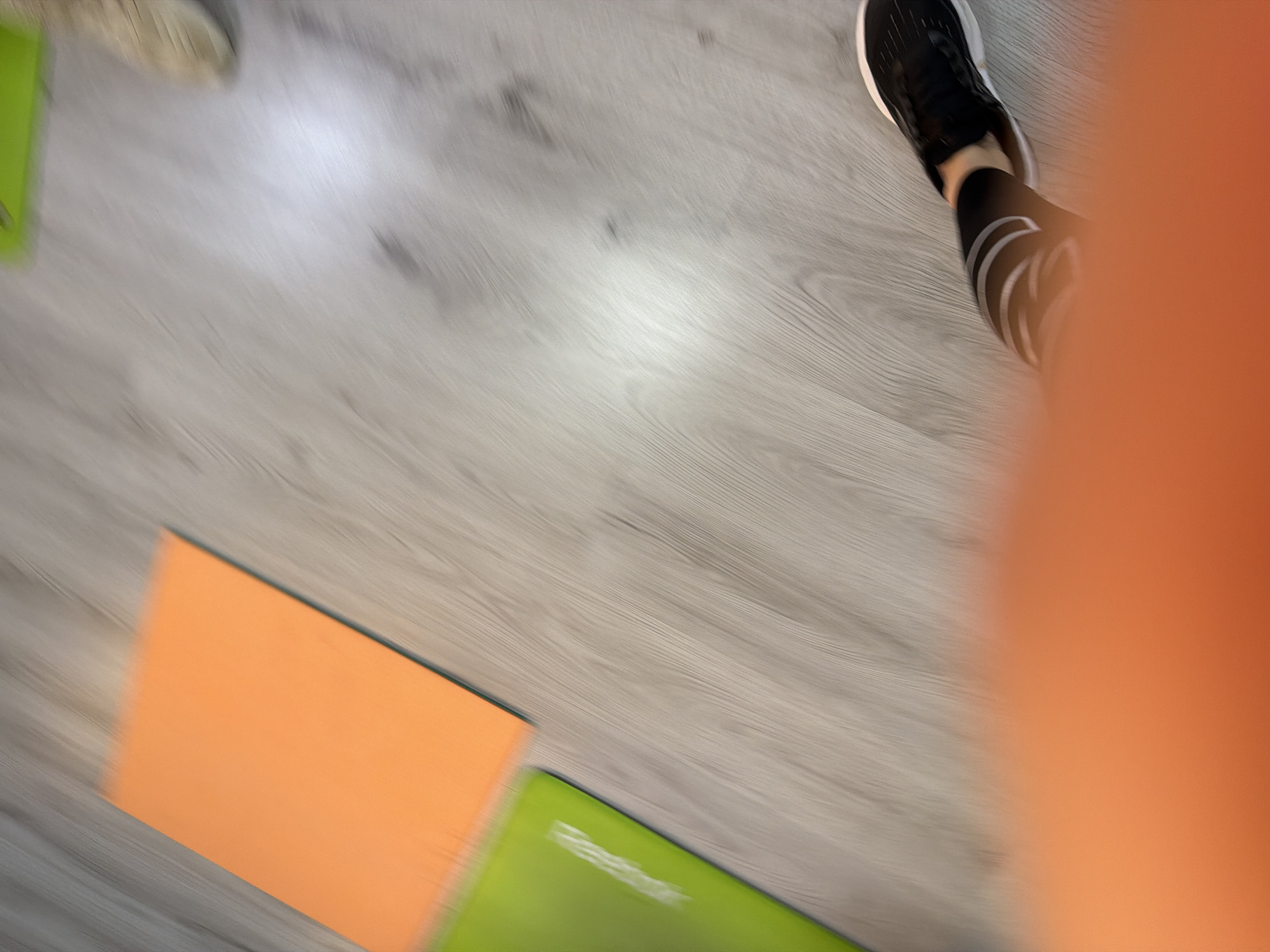 This image captures an indoor scene, likely a fitness studio or gym in Izhevsk, Russia, characterized by significant motion blur across the entire frame. The light-colored wooden or laminate floor serves as the primary background. In the upper right quadrant, a person's lower right leg and foot are visible, clad in black athletic pants featuring white stripes or a pattern, and a black athletic shoe with a white sole. Another blurred, lighter-colored foot or shoe is partially visible in the upper left corner.
On the floor in the lower left, an orange rectangular mat or board is positioned, with a green mat partially visible beneath and to its right. Blurred white text, illegible due to the motion, is present on the green mat. A large, out-of-focus orange blur dominates the right side of the image, potentially a part of the person's clothing or another object caught in motion. The overall impression is one of activity or movement, possibly during an exercise session, under indoor lighting.