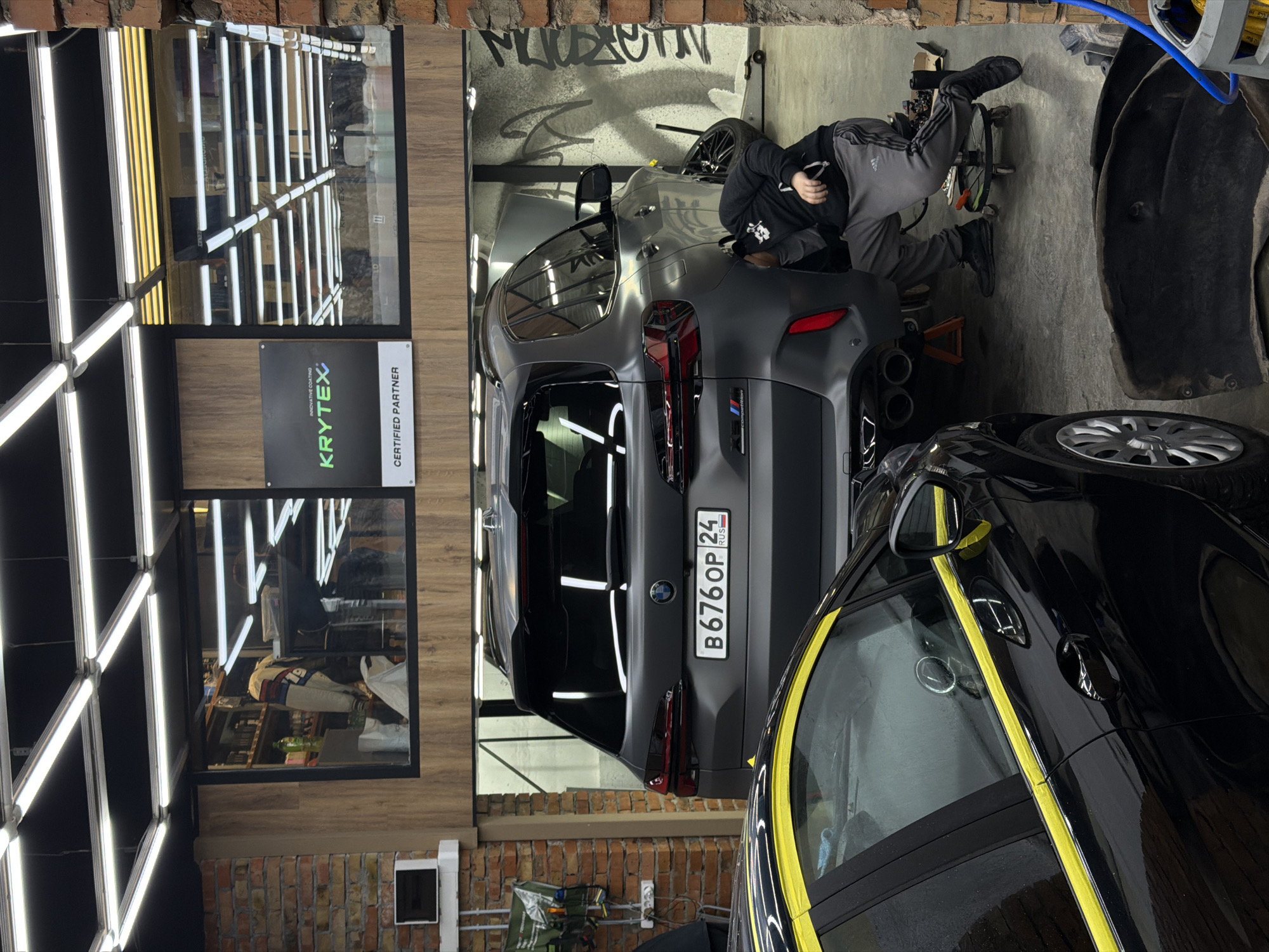 A photograph captures a scene within what appears to be an auto detailing or repair shop in Krasnoyarsk, Russia. The image is oriented vertically. A matte gray BMW X5 M Competition is positioned prominently. A man in a black hoodie and gray sweatpants is working on the rear of the car. A black vehicle, partially visible and covered with yellow tape, is situated in the foreground.
The license plate of the BMW reads "B676OP 24 RUS," indicating its origin. The shop is well-lit with several rows of fluorescent lights. A window displays a "KRYTEX" sign, suggesting a partnership with a coating product company. Brick walls are visible in the foreground and background. Spray paint graffiti marks the wall behind the gray BMW. Auto repair equipment is scattered around the workspace. The scene gives the impression of active car maintenance or detailing.