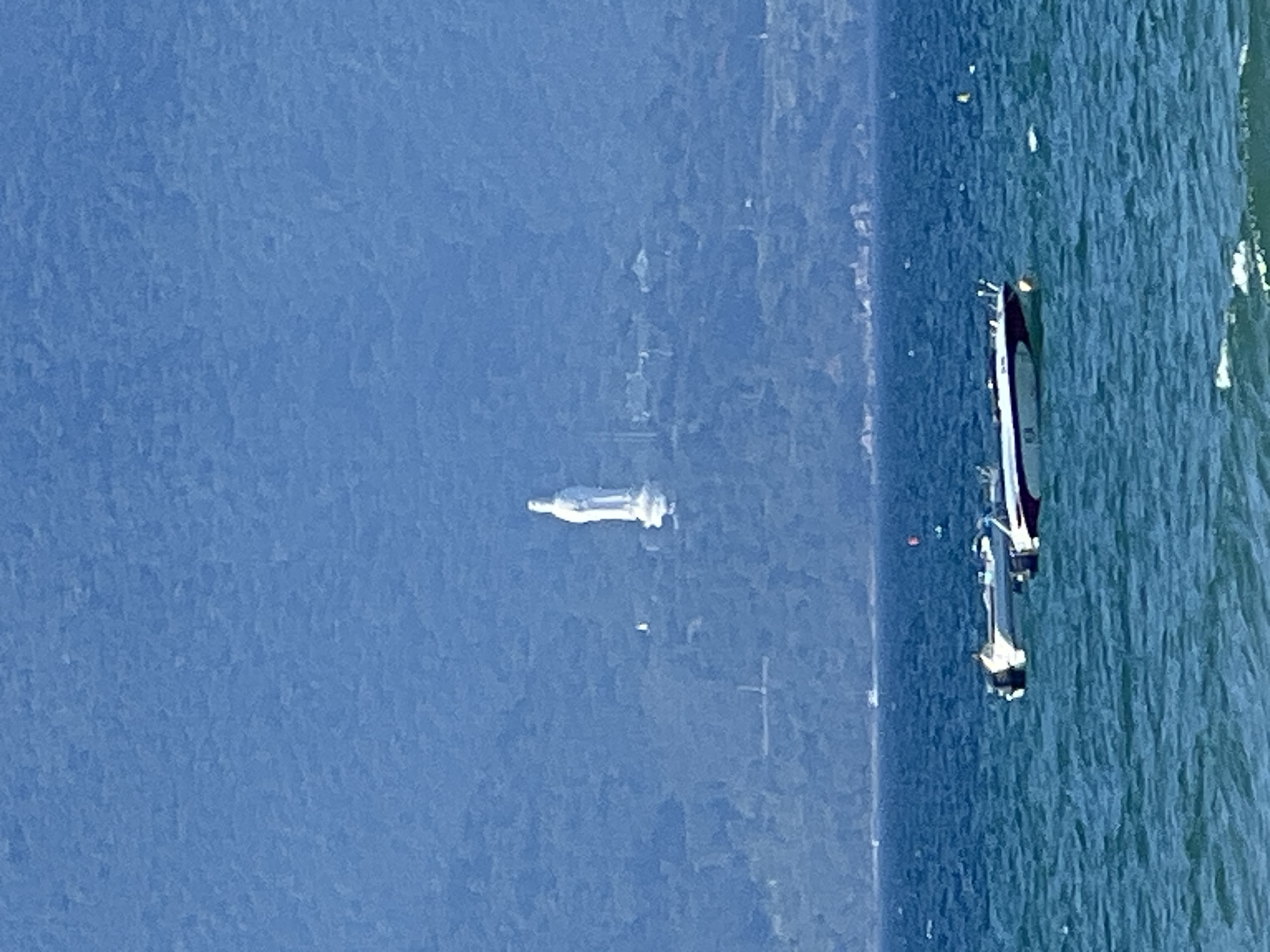 A wide shot captures a serene seascape with a prominent white statue situated on a verdant hillside in the distance. The statue, a significant landmark in Da Nang, Vietnam, is visible atop Son Tra Peninsula, overlooking the South China Sea. Two speedboats are anchored in the foreground, bobbing on the gently rippling blue water. The sky above is a clear, bright blue, with a vast, lush green mountain range serving as a backdrop.