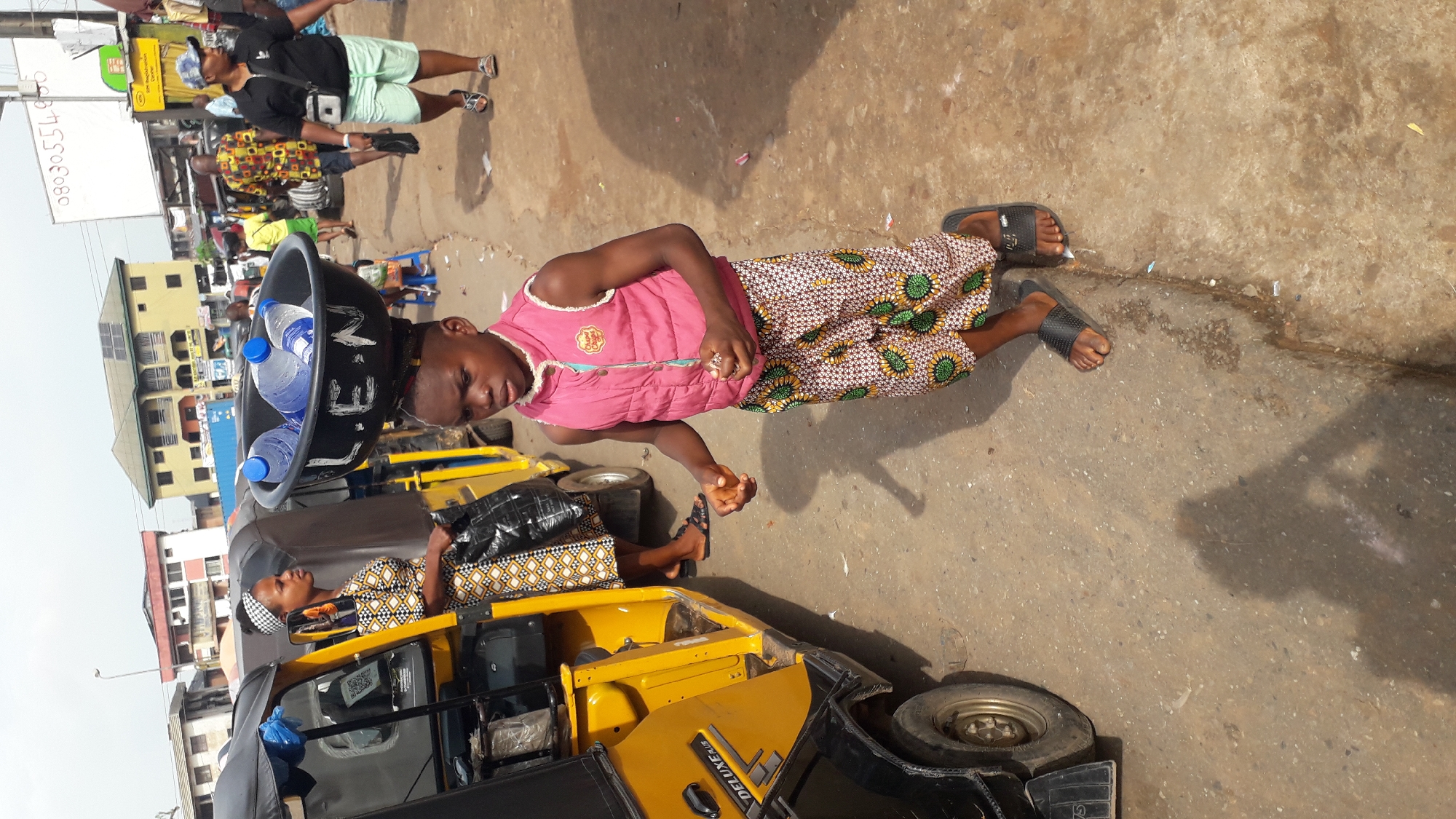 In Nkpor, Nigeria, a young boy, likely under ten years old, is seen carrying a black basin filled with several plastic bottles of water on his head. He wears a pink vest over a patterned skirt and black slide sandals. To his left, a woman in a patterned dress and headscarf stands next to a yellow tricycle-style auto-rickshaw. The scene unfolds on a dusty, paved street, with other individuals and buildings visible in the background, suggesting a bustling market or street environment.