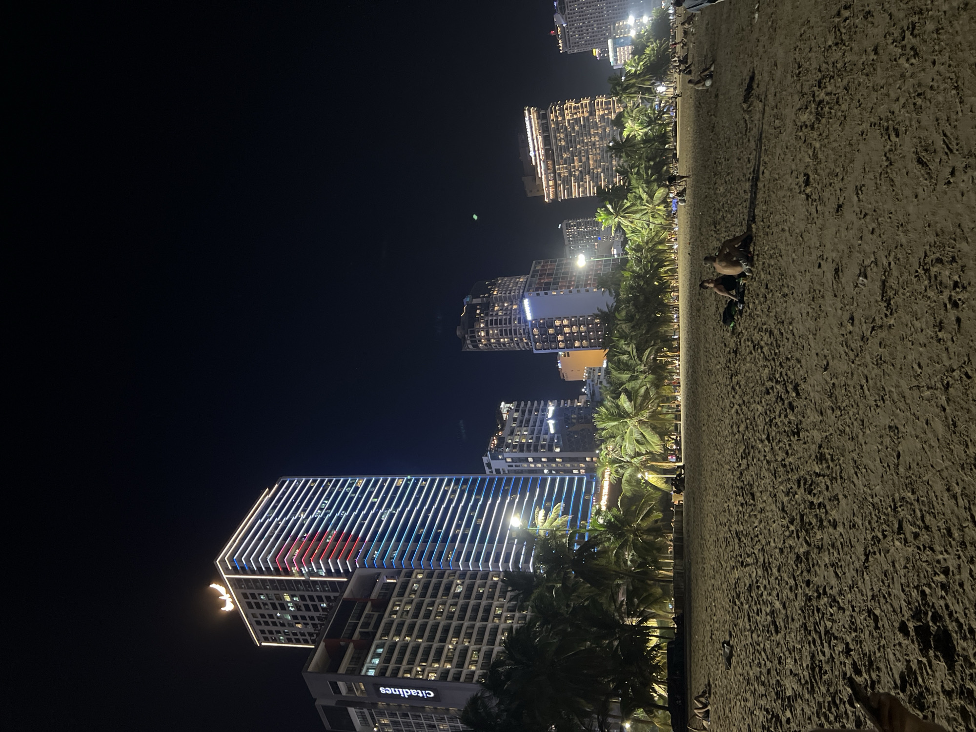 The image depicts a nighttime scene on a sandy beach in Nha Trang, Vietnam. In the background, a row of modern, illuminated skyscrapers lines the coast, with palm trees in the foreground casting shadows on the sand. Several people are scattered across the beach, some sitting and relaxing. One group of two individuals sits together on a blanket, facing away from the camera. The overall atmosphere is calm and leisurely.