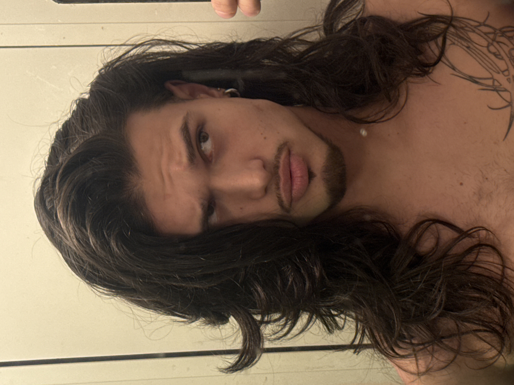 A young man with long, dark, wavy hair and a goatee is pictured from the chest up. He is looking to his right with a slightly furrowed brow and a pout. He is wearing a silver hoop earring in his left ear and a small white pearl on his chest. A portion of a tattoo is visible on his right chest. The background appears to be a wall in a dimly lit room, possibly a bathroom, with a vertical light fixture on the left. The image is a selfie taken with a phone, indicated by the reflection of a hand holding the phone in the upper right corner. The location in Sofia, Bulgaria, is not discernable from the image itself.