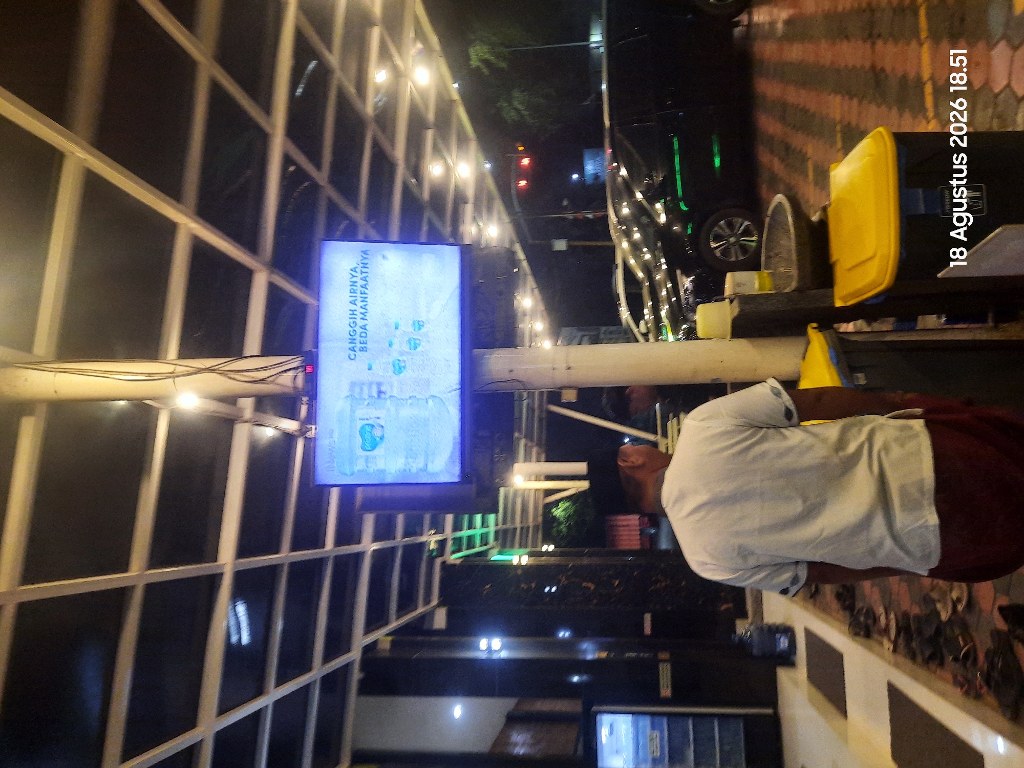 The image shows a man from the back, wearing a white shirt and a maroon sarong, standing in front of a large television screen. The screen displays an advertisement for water bottles, with text in Indonesian that translates to "Sophisticated, its air is beneficial." The scene appears to be outdoors, possibly in a public area like a rest stop or a market, given the presence of trash cans and shoes lined up on the ground. In the background, cars are parked, and trees are visible. The overall atmosphere is that of an evening or night setting, with artificial lighting illuminating the area. The date stamp in the lower right corner indicates "18 Agustus 2026 18.51", which translates to August 18, 2026, 6:51 PM. The location is identified as Sukokerto, Indonesia.