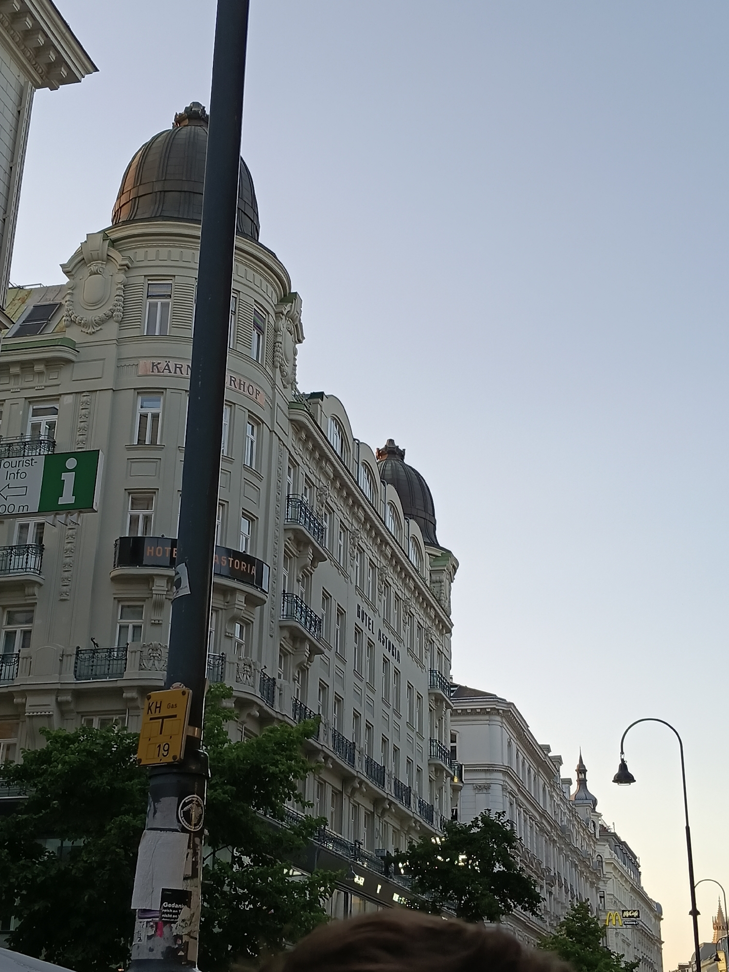 This image captures a view of the Kärntnerhof Hotel in Vienna, Austria, situated on a street lined with ornate architecture. The Kärntnerhof Hotel, a prominent building, features a distinctive dome and classical architectural details. A street lamp with a curved arm and a dark lampshade stands prominently in the foreground, slightly obscuring parts of the building. Several signs are visible, including one for "Tourist-Info" with an arrow pointing to the left and a distance of "100 m," and another with "HOTEL ASTORIA" emblazoned on it. A street sign with "KH Gas T 19" is also attached to the lamp post. Trees with lush green foliage are situated in front of the buildings, and the sky above is clear and pale. The overall scene suggests a bright day in a European city.