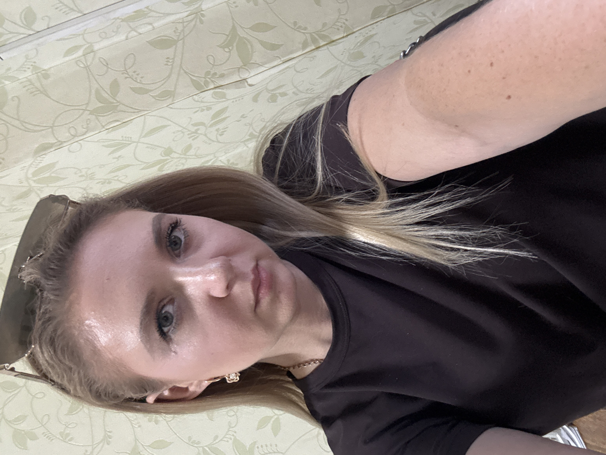 A young blonde woman with blue eyes is pictured from the chest up, looking directly at the camera. She is wearing a black t-shirt and gold hoop earrings with flower charms. Sunglasses are perched on her head, and a thin gold chain necklace is visible. Her blonde hair is parted in the middle and falls past her shoulders. The background consists of a patterned wallpaper in a pale green color. The lighting suggests an indoor environment.
