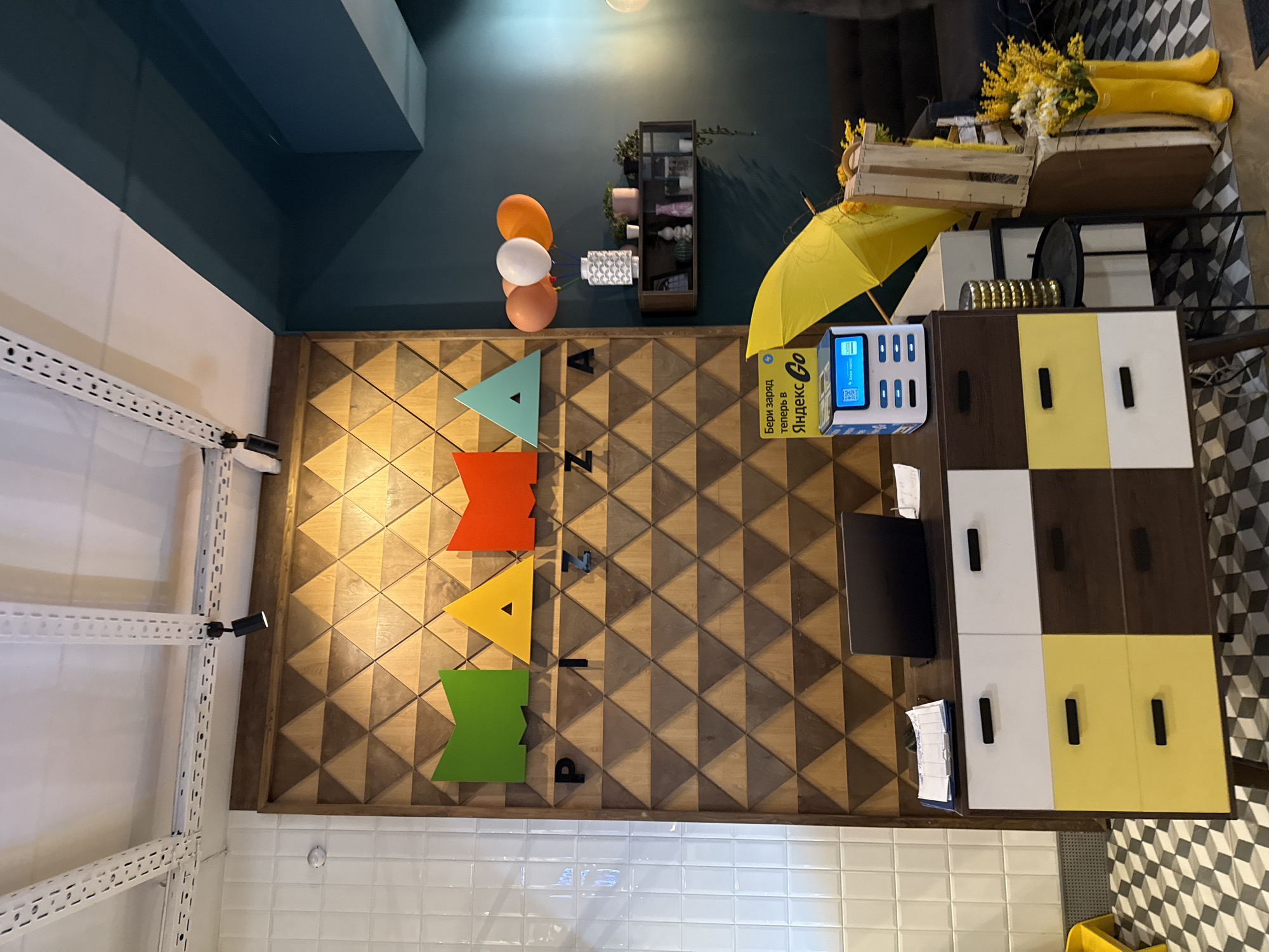 The image presents an indoor scene, likely a commercial space or eatery. The wall features a decorative wooden panel displaying the word "PIZZA" vertically, with each letter rendered in a different color (teal, orange, yellow, and green). Above the panel, balloons in orange and white are visible. A small, dark shelf unit with decorative items is mounted on the wall.
To the right of the panel, a yellow umbrella is positioned next to a phone charging station branded "Яндекс Go" (Yandex Go). A desk with a brown and white cabinet adjoins the charging station. Papers and a black folder are present on the desk's surface.
The walls are painted in two-tone, white in the lower section and teal above. A shelving system is mounted on the white section of the wall. The floor is tiled in white and a patterned black and white tile. The setting potentially depicts an interior of a pizza restaurant in Izhevsk, Russia, given the Cyrillic branding on the charging station.