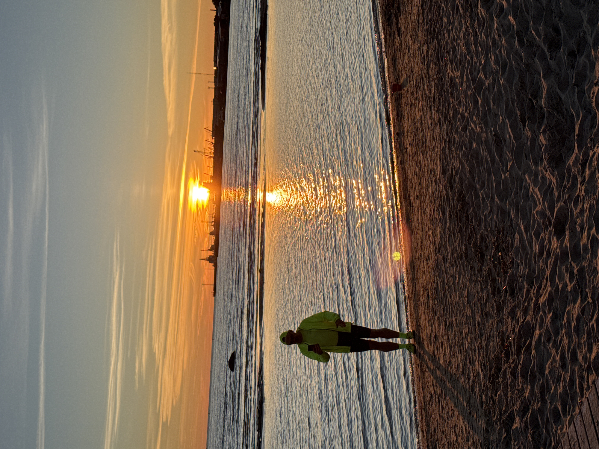 A person wearing a neon yellow jacket and black shorts is standing on a sandy beach next to a calm body of water. The sun is setting, casting a warm orange and yellow glow across the sky and reflecting off the water's surface. In the distance, a port with cranes is visible on the horizon. The sandy beach has ripples and texture from the wind. The image suggests a peaceful and serene moment, possibly during an evening run or walk. The time of day is clearly sunset. The location is identified as Tallinn, Estonia. There is no visible text in the image.