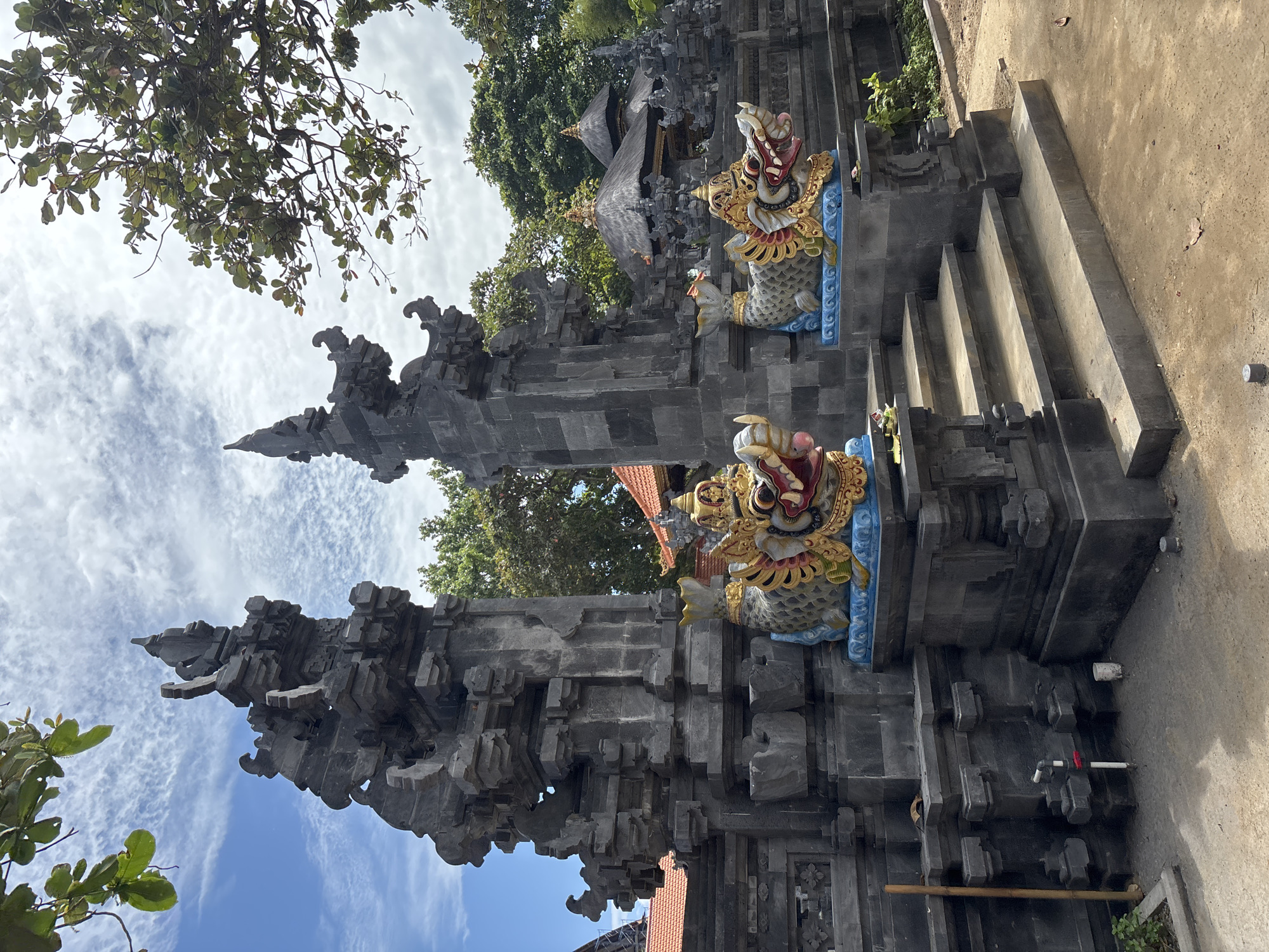 This is a full-shot, eye-level photograph of a Balinese temple entrance in Kuta Selatan, Indonesia. The temple is made of dark grey stone with intricate carvings. Flanking the entrance are two decorative statues of dragon-like creatures, brightly colored with red, yellow, and blue accents. These statues sit on blue bases, and yellow, leafy decorations adorn their heads. A set of stone stairs leads up to the temple entrance, with two steps visible in the foreground. Behind the temple entrance, a traditional Balinese building with a thatched roof can be seen. Lush green trees surround the temple, and the sky above is a mix of blue and white clouds. The ground in front of the temple is sandy and bare, with a few fallen leaves scattered about. A bamboo pole stands vertically to the left of the stairs, and a white pipe with a red valve is visible near the base of the temple wall.