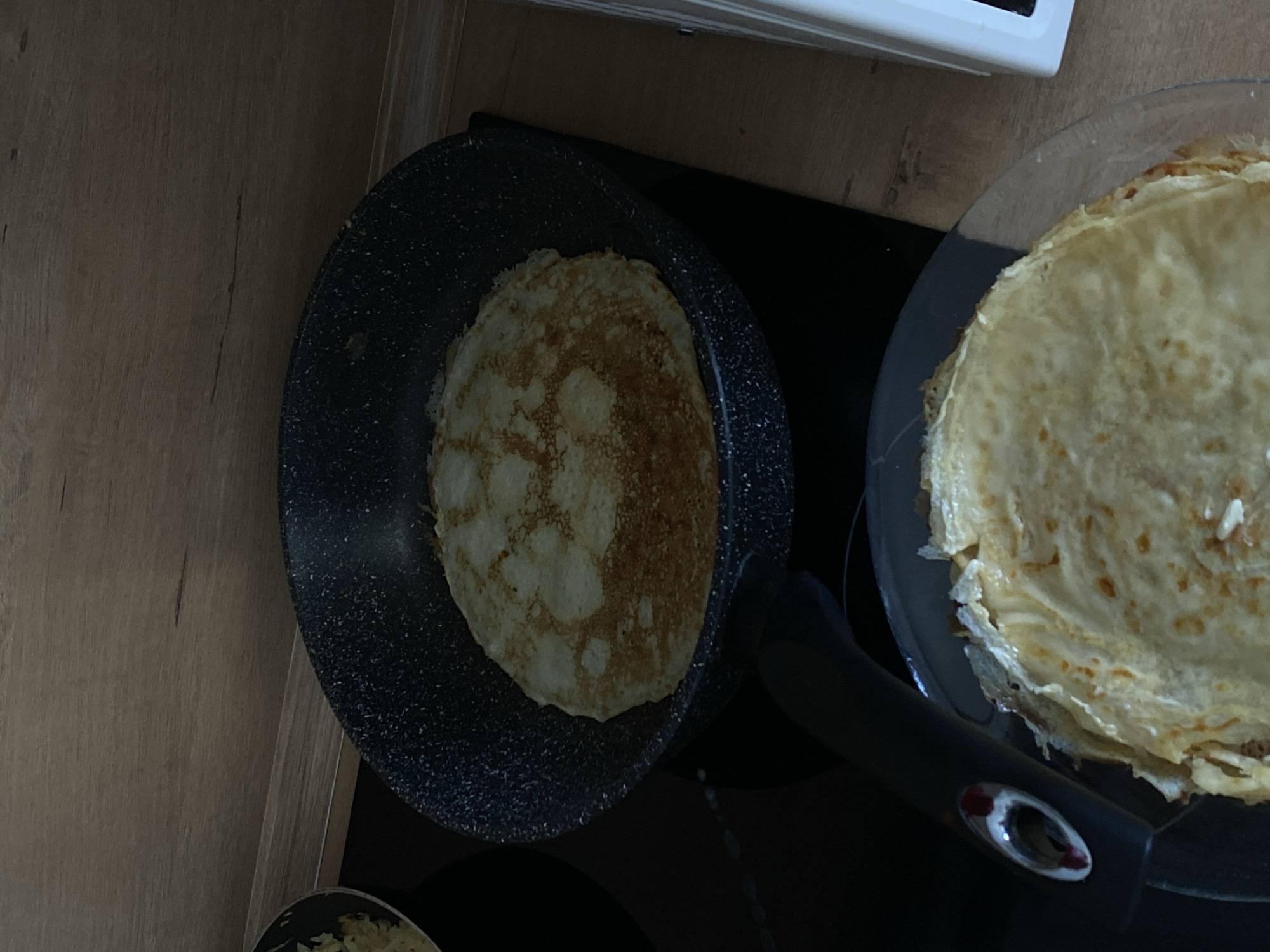 A top-down shot shows a black speckled frying pan with a cooked crepe in it, next to a clear glass plate holding a stack of several cooked crepes. The scene appears to be in a kitchen, likely in Kazan, Russia, given the user's request. The frying pan and plate rest on a black cooktop surface, adjacent to a light-colored wooden countertop. A white appliance is partially visible in the upper portion of the image.