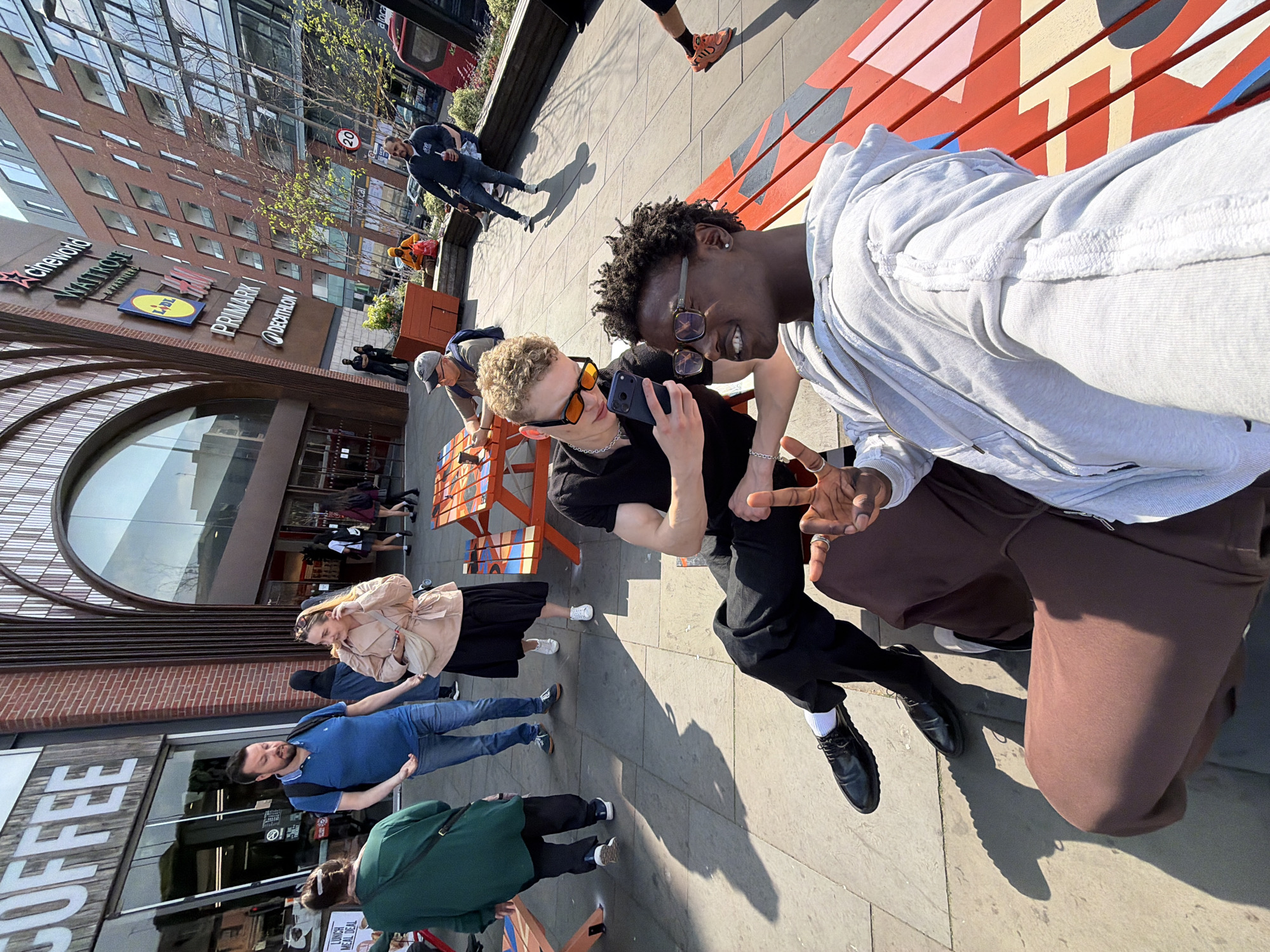 This image captures a vibrant urban scene in Watford, London, United Kingdom, under bright daylight with strong shadows indicating a sunny day. The primary subjects are two young men in the foreground, seated on or near a colorful, geometrically patterned wooden bench. The man on the right, with dark curly hair and glasses, wears a light grey hooded top and brown trousers, smiling and making a hand gesture. Beside him, a man with blonde curly hair and orange-tinted sunglasses, dressed in a black t-shirt and black trousers, holds a smartphone (likely an iPhone) pointed towards them, suggesting they are taking a selfie or engaging with the device. Both appear cheerful.
In the mid-ground and background, other pedestrians populate the paved street. Notable individuals include a man in a blue polo shirt and jeans with a backpack, and a woman in a light-colored jacket and dark skirt. The setting is characterized by large commercial buildings, one featuring prominent signage for various retail outlets.
Visible text on the buildings includes "CINEWORLD" with its red star logo, "WATFORD," "LIDL" (blue and yellow logo), "PRIMARK" (pink text), and "DECATHLON" (blue text). Further down the street, "COFFEE" is visible in large white letters on a building facade, along with a smaller sign for "LUNCH MEAL DEAL." A small red road sign with "20" is also visible in the distance, indicating a speed limit. The overall scene depicts a bustling commercial area on a pleasant day.