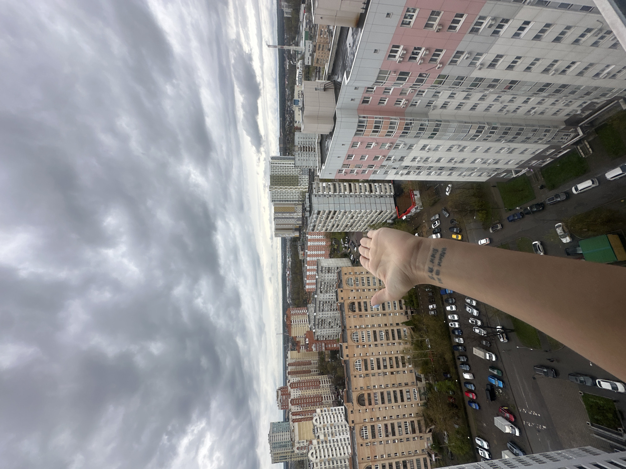 A high-angle, eye-level shot depicts a section of Khimki, Russia, under an overcast sky. Multiple tall apartment buildings, varying in color and design, dominate the view. Cars are parked along the roads between the buildings. An arm and hand with a tattoo are visible in the foreground, extending into the scene.