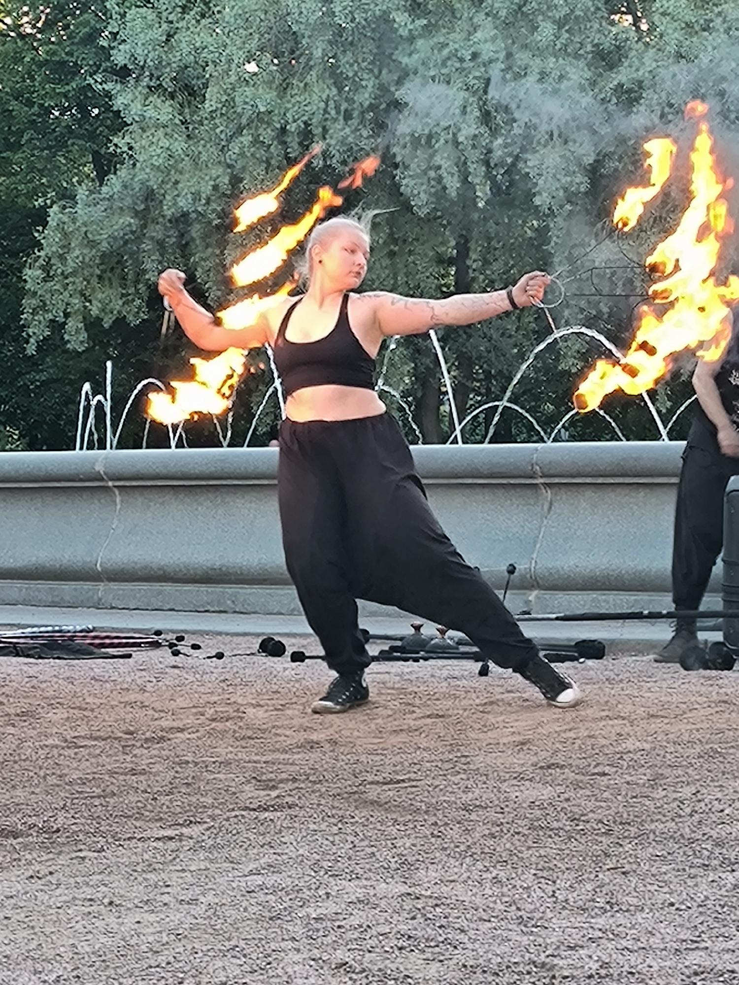 A woman with blonde hair pulled back is performing with fire props in front of a fountain and trees. She is wearing a black sports bra and black, loose-fitting pants. She is holding two fire props, one in each hand, which are lit and producing flames. Her body is angled to her left, and her arms are extended, one forward and one to the side. In the background, a fountain sprays water upwards. Behind the fountain, there are lush green trees. On the ground in front of the woman, there are several objects that appear to be props for fire performance, including hoops and sticks with weighted ends. Another person, partially visible on the right side of the frame, is also wearing black pants and appears to be involved in the performance. The ground is covered in a reddish-brown gravelly material. The scene appears to be taking place outdoors in a park or public square.