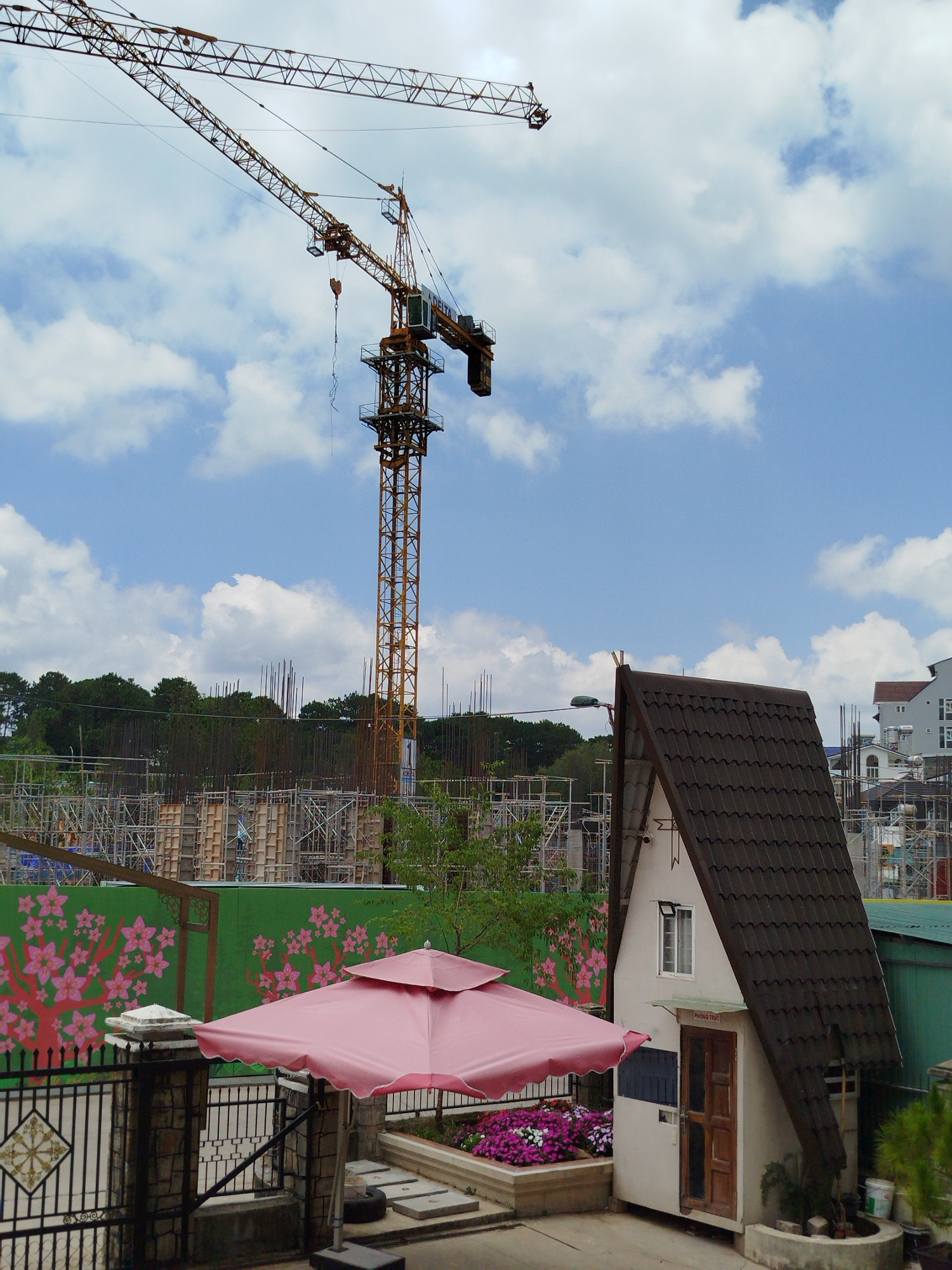 The image depicts a scene in Da Lat, Vietnam, characterized by a mix of construction and residential elements. A tall yellow construction crane dominates the skyline against a backdrop of a partly cloudy sky. Beneath the crane, there is a construction site with steel rebar and scaffolding visible. In the foreground, there is a small, A-frame-style building with a dark tiled roof, situated beside a green wall adorned with pink flower murals. A pink umbrella provides shade over a flowerbed filled with purple and white flowers. A black wrought-iron fence partially obscures the lower left corner of the image. In the distance, behind the construction site and green wall, there is a line of trees and additional buildings, indicating a residential area.