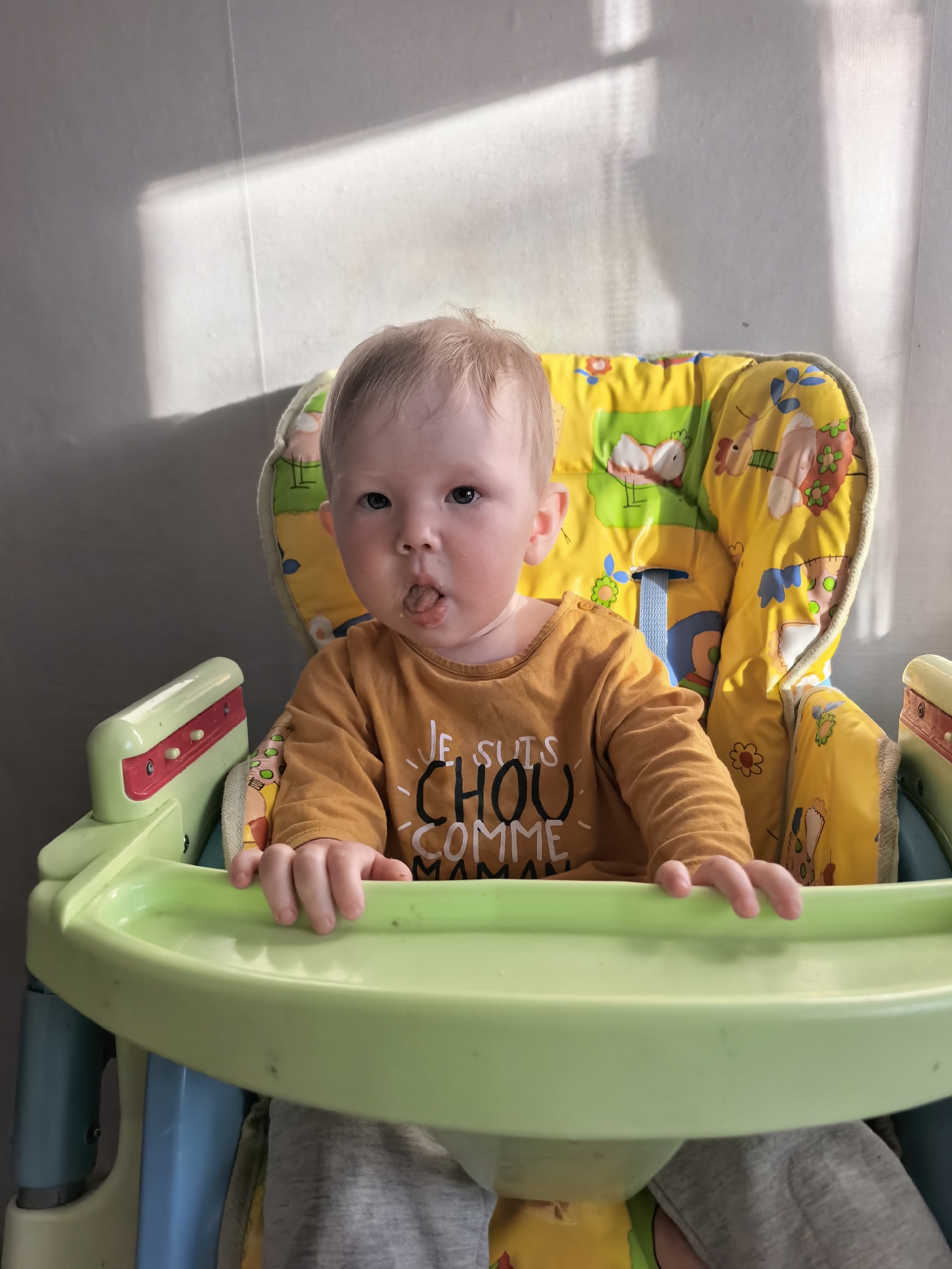 A fair-skinned toddler is sitting in a colorful highchair, appearing to be indoors. The background is a plain wall with shadows cast by sunlight. The toddler is wearing an orange long-sleeved shirt. The highchair has a yellow padded seat with various cartoon animal patterns. The tray of the highchair is green, and the legs are a combination of blue and green. The toddler has light blonde hair and is sticking their tongue out slightly. There is no indication of the location being Sterlitamak, Russia, within the visual context of this image.