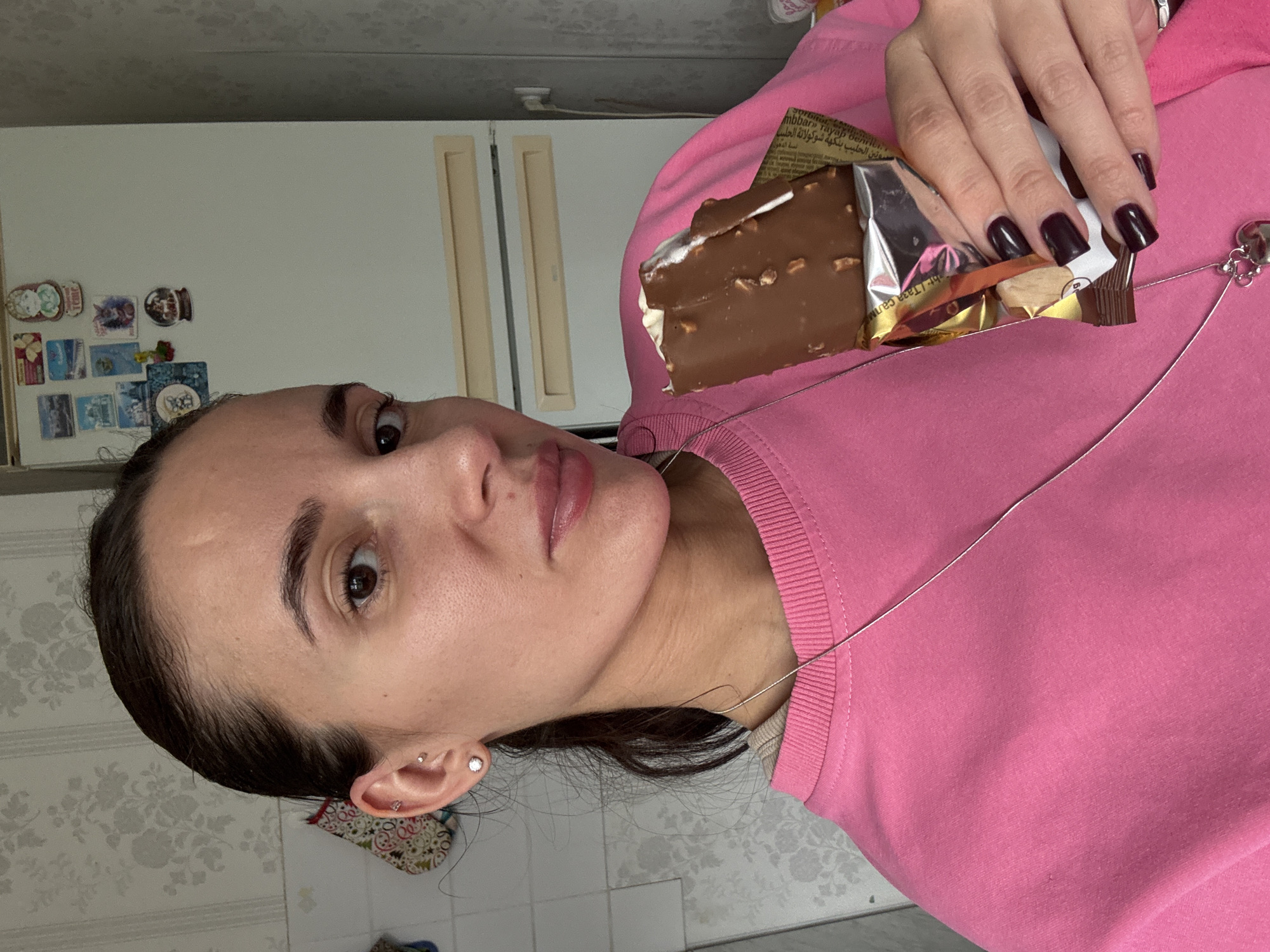 A woman with dark hair pulled back is holding a partially eaten chocolate bar in her hand. She is wearing a pink sweatshirt and a silver necklace. Her fingernails are painted dark purple. She appears to be in a kitchen, with a refrigerator visible in the background, decorated with magnets. The location cannot be ascertained to be Chaikovsky, Russia from the provided information.