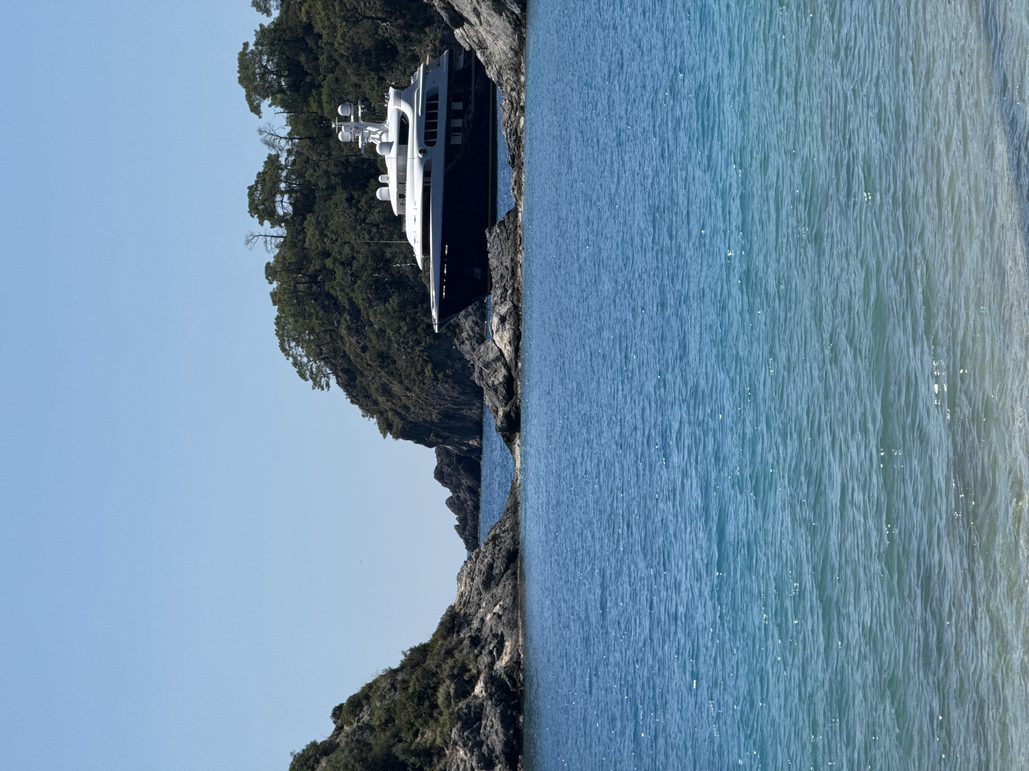 The image shows a large yacht docked near a rocky coastline with lush green vegetation. The yacht is predominantly white with dark accents and several levels, suggesting a luxury vessel. The water is a mix of blue and green hues, indicating shallow depths near the shore. The coastline features jagged rocks and dense foliage. The sky is clear and light blue. The location is likely in a coastal area near Fethiye, Turkey, due to the characteristic turquoise waters and mountainous, forested landscapes often seen in the region.