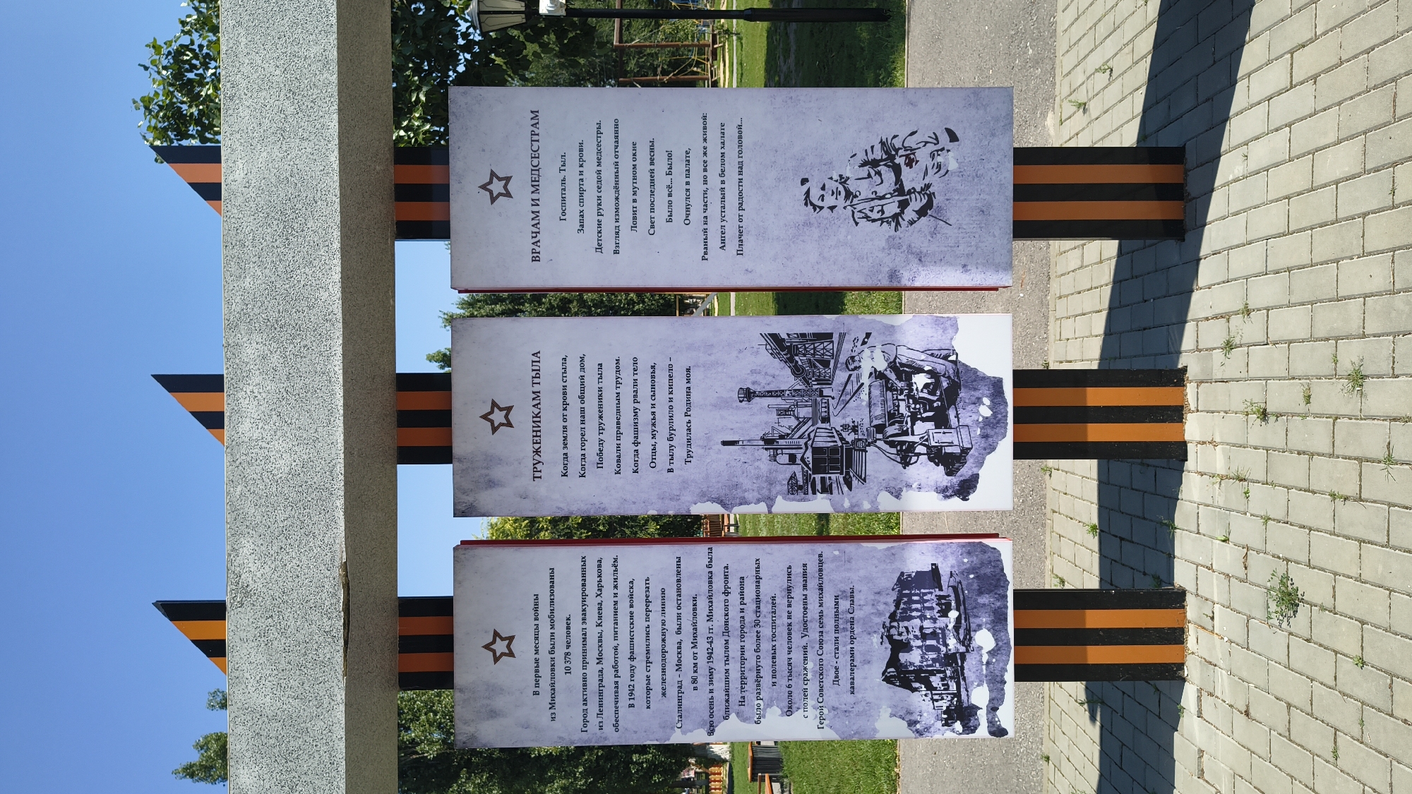 Three informational panels stand in an outdoor setting, likely a park or public square in Mikhaylovka, Russia, given the context of the text on the panels. The panels are vertically oriented and supported by black and orange striped posts, reminiscent of the colors of the Order of St. George ribbon, a symbol of military valor in Russia. Each panel features a red border and a white background with text and an illustration.
The left panel, titled "В первые месяцы войны из Михайловки были мобилизованы 10 378 человек," details the mobilization of over 10,000 individuals from Mikhaylovka at the start of the war, their role in evacuating and supplying Leningrad, Moscow, and Kharkov, and their contribution to the construction of a railway line. The accompanying illustration depicts a building, possibly a factory or administrative structure.
The central panel, titled "ТРУЖЕНИКАМ ТЫЛА," honors the home front workers. The text describes their sacrifices, the hardships they endured, and their role in supporting the war effort. The illustration shows a factory with smoking chimneys and figures working, conveying an industrial and wartime atmosphere.
The right panel, titled "ВРАЧАМ И МЕДСЕСТРАМ," is dedicated to doctors and nurses. The text describes the grim realities of a hospital during wartime, the dedication of medical personnel, and the emotional toll of their work. The illustration shows a female nurse in uniform, looking contemplative, with a faint outline of medical equipment or a patient behind her.
The panels are arranged in a row, facing the viewer. Behind them, a paved walkway made of rectangular grey bricks is visible. The background consists of green trees and a clear blue sky. The overall scene suggests a memorial or educational display commemorating the sacrifices and contributions made during wartime.