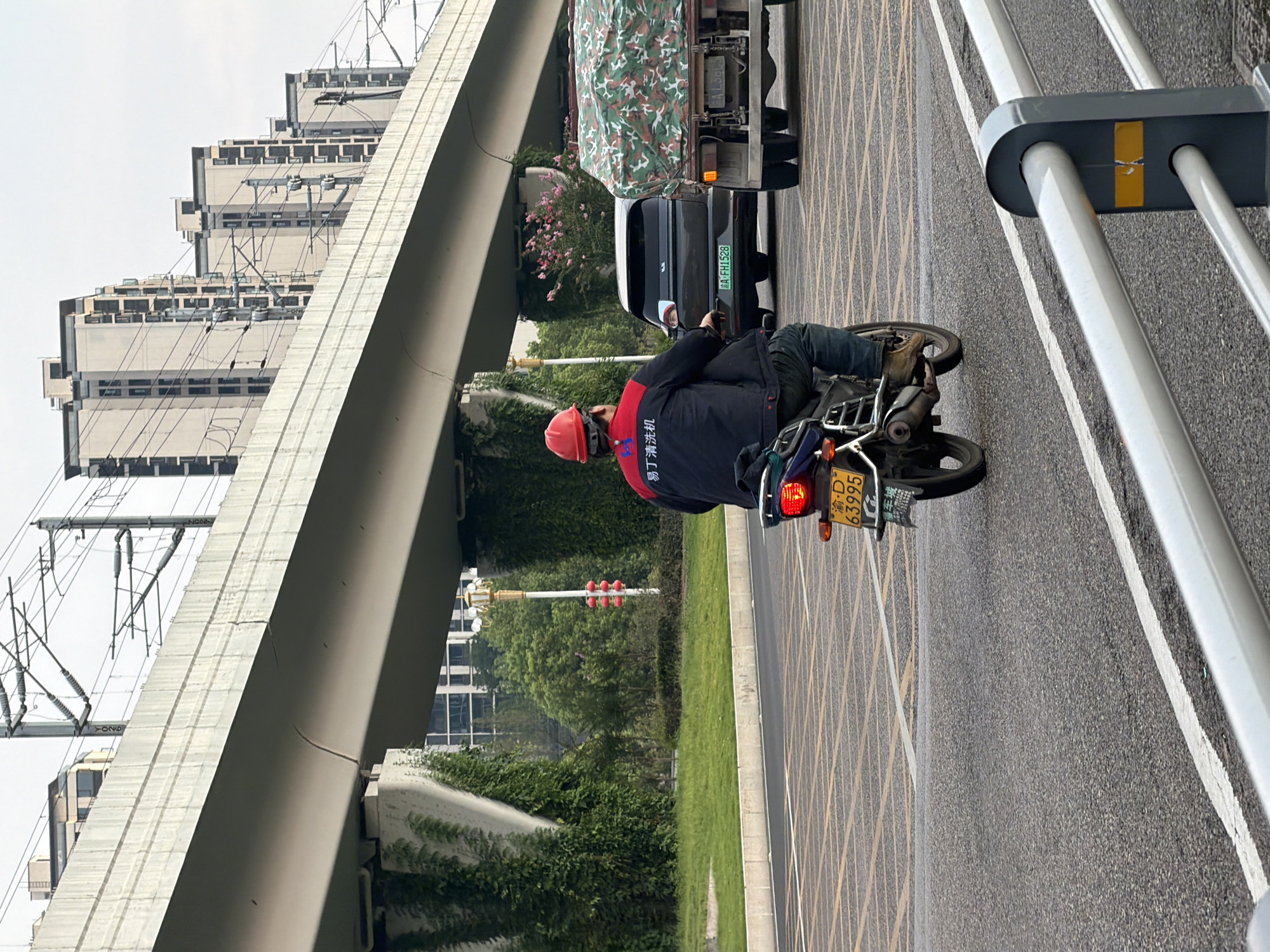 In Jiangjin, China, a man wearing a red hard hat and a dark jacket with Chinese lettering on the back is riding a motorcycle. The motorcycle has a yellow license plate with Chinese characters and the numbers "63995". The man is on an asphalt road with yellow crosswalk markings. In the background, a large elevated concrete structure, possibly a bridge or overpass, runs horizontally across the image. This structure is supported by concrete pillars covered in green ivy. Beyond the overpass, several multi-story buildings are visible, with a clear sky above. A dark-colored car and a truck with a camouflage-covered load are also on the road.