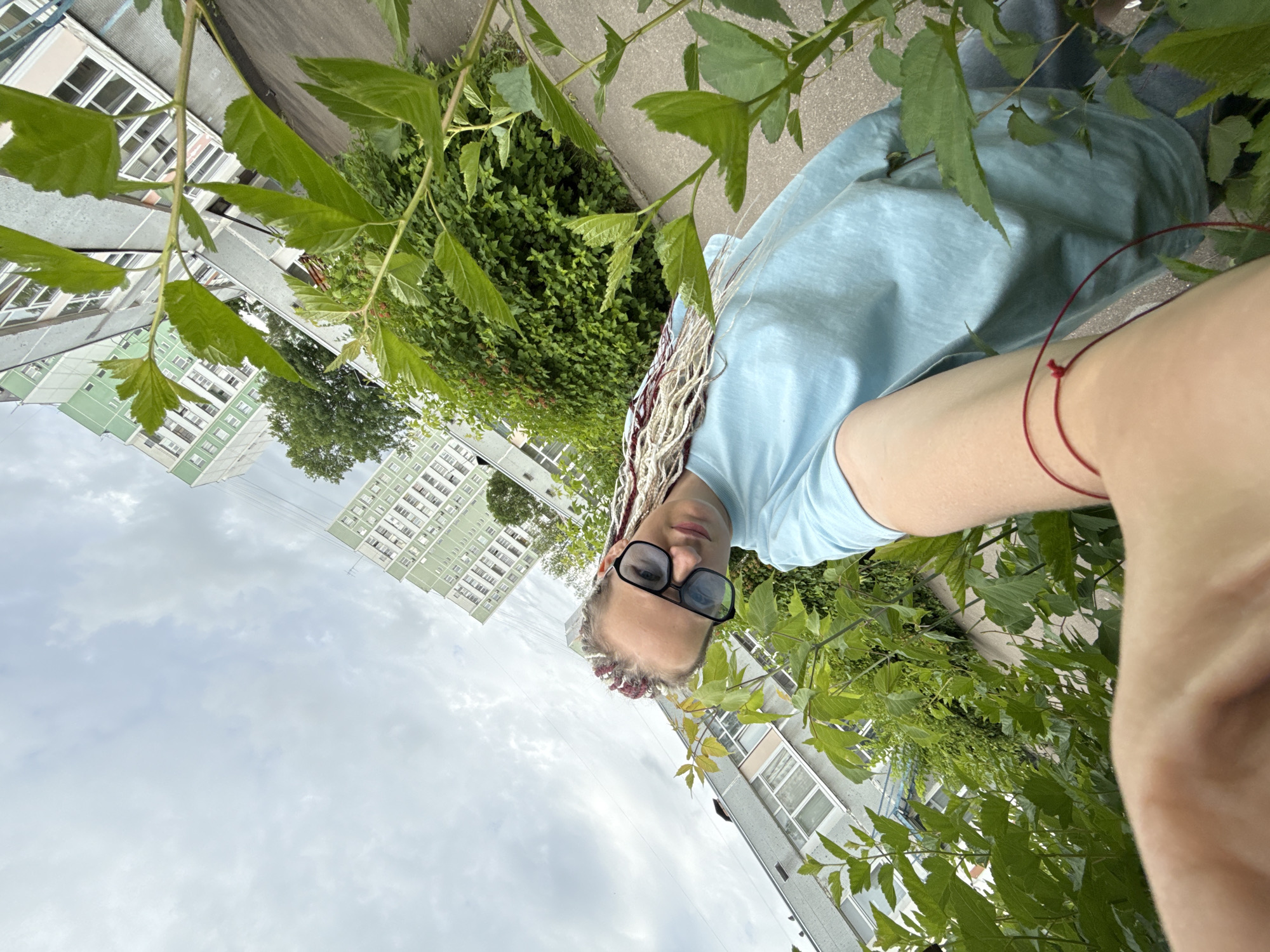 A person with blonde braids, some of which have a pinkish hue, wearing black-rimmed glasses and a light blue t-shirt, is holding a phone to take a selfie. They are outdoors, with lush green foliage in the foreground and to the sides. In the background, multiple apartment buildings are visible, some with light green facades and others with a beige and grey exterior. The sky is overcast with clouds. The scene appears to be a residential area in Moscow, Russia, likely a courtyard or common area between buildings. The perspective is from a low angle, looking up towards the person and the buildings. A thin red string is tied around the person's left wrist.
