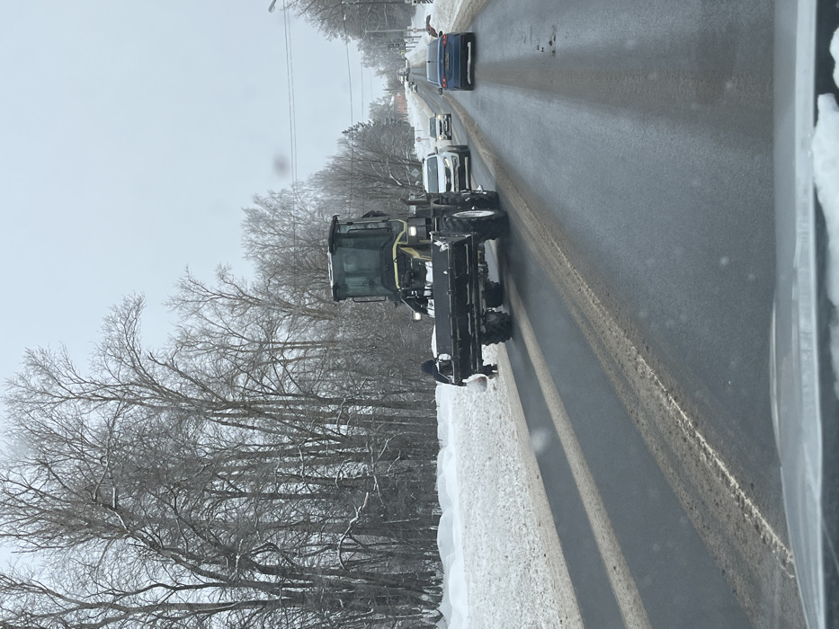 This image depicts a winter roadside scene in Ширяево, Russia, captured from inside a vehicle. A large, yellow and black front-end loader or tractor with a wide black snow shovel attachment is positioned prominently on the right side of the asphalt road. Its headlights are illuminated. To the immediate left of the tractor, on the snow-covered verge, a person wearing dark winter clothing stands, holding a light-colored bag. Another similar bag is visible on the snow near their feet, suggesting an interaction with the ground or the tractor. The road surface is dark, wet, and shows patches of slush and dirt, with cleared tire tracks. A snowbank lines the left shoulder of the road, behind which stands a dense grouping of bare deciduous trees dusted with snow. In the distant right, several passenger vehicles, including a white SUV and a dark blue sedan, are traveling away from the camera. Overhead utility lines and poles extend along the road, with vague structures visible in the far background under an overcast sky.