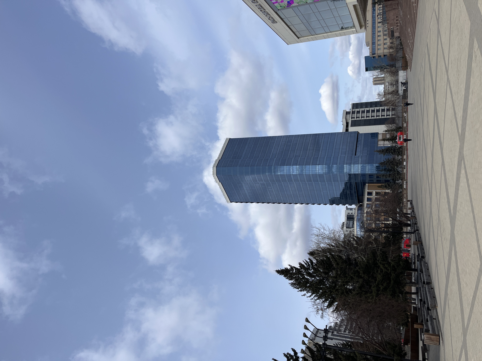 The image depicts a view in Krasnoyarsk, Russia, looking upwards towards the sky and surrounding architecture. A modern, glass-facade skyscraper dominates the center of the frame, reflecting the sky and clouds. To the right, a paved area with a geometric pattern is visible. Trees are interspersed among the buildings. The sky features a mix of blue and clouds. A few people are seen on the paved area in the distance.