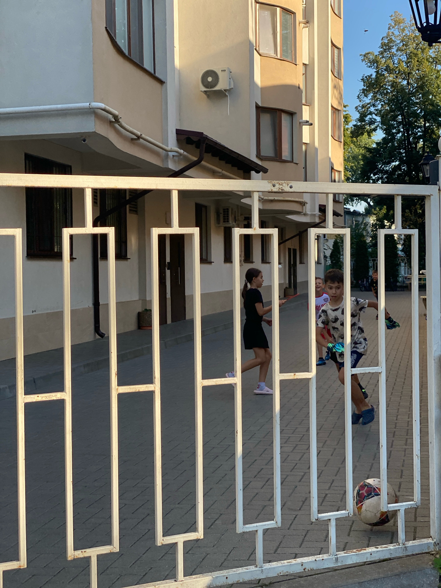 This image depicts a scene outside a modern building in Chișinău, Moldova, viewed through a white metal gate. In the foreground, the gate's bars create a visual obstruction, with the lower portion of a multi-colored soccer ball visible resting on the pavement. Beyond the gate, on a paved walkway, three children are engaged in activity. A girl in a black dress and pink socks is in motion, facing left. To her right, a boy wearing a patterned t-shirt and dark shorts is in the process of kicking the soccer ball. Another boy, in the background and slightly out of focus, appears to be laughing. The building behind them has light-colored facades, with visible windows and architectural features like a white pipe and an air conditioning unit. Trees and a lamppost are present in the background, suggesting an urban or residential setting on a clear day.