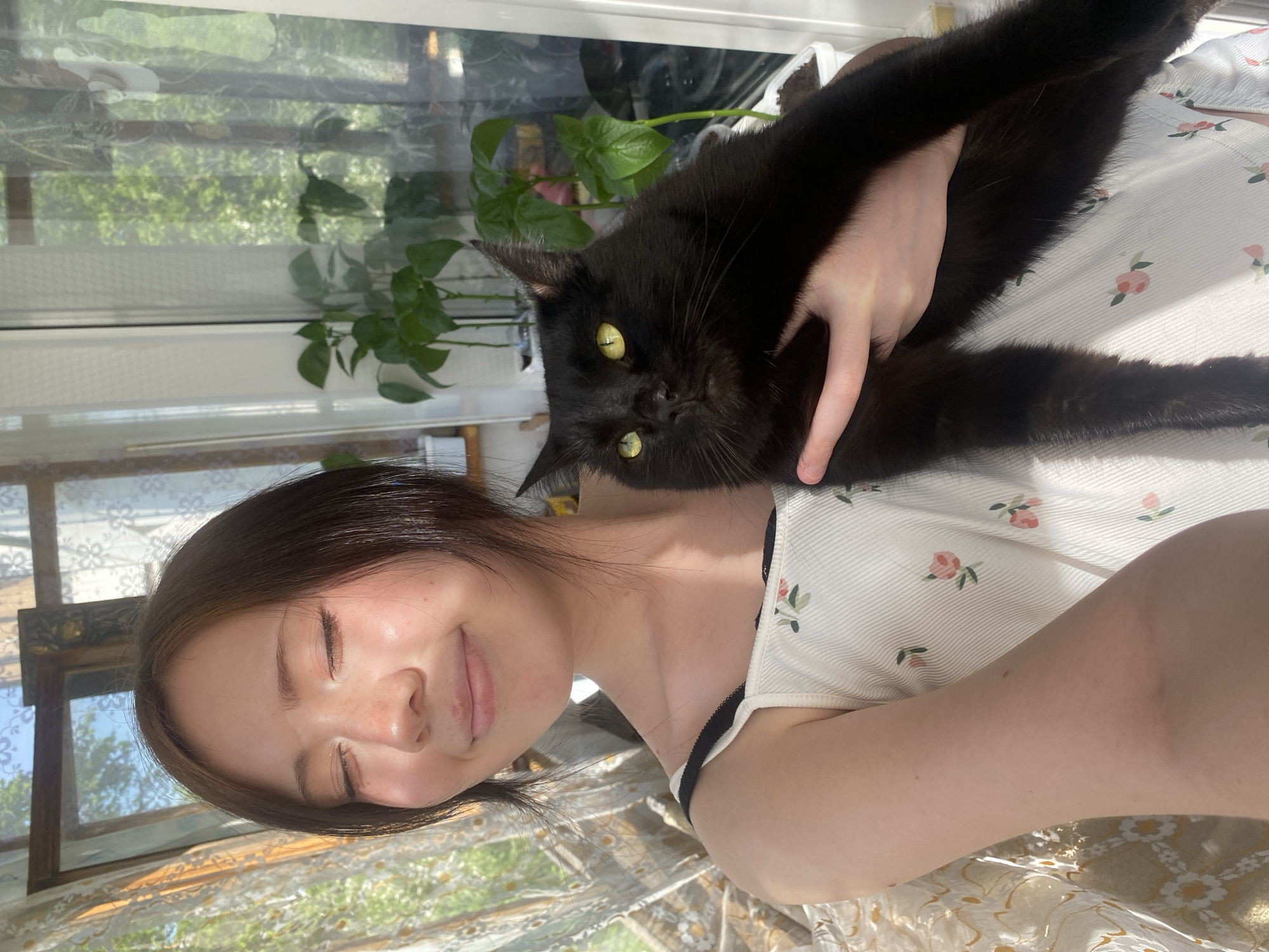 A woman is holding a black cat. The woman has brown hair, light skin and freckles. The cat has yellow eyes. They are inside near a window with a view of trees. A green leafy plant is visible in front of the window. The woman is wearing a light-colored top with a floral pattern. The image is taken in a well-lit area. The location is not identifiable as Unknown, Unknown based on the provided image.