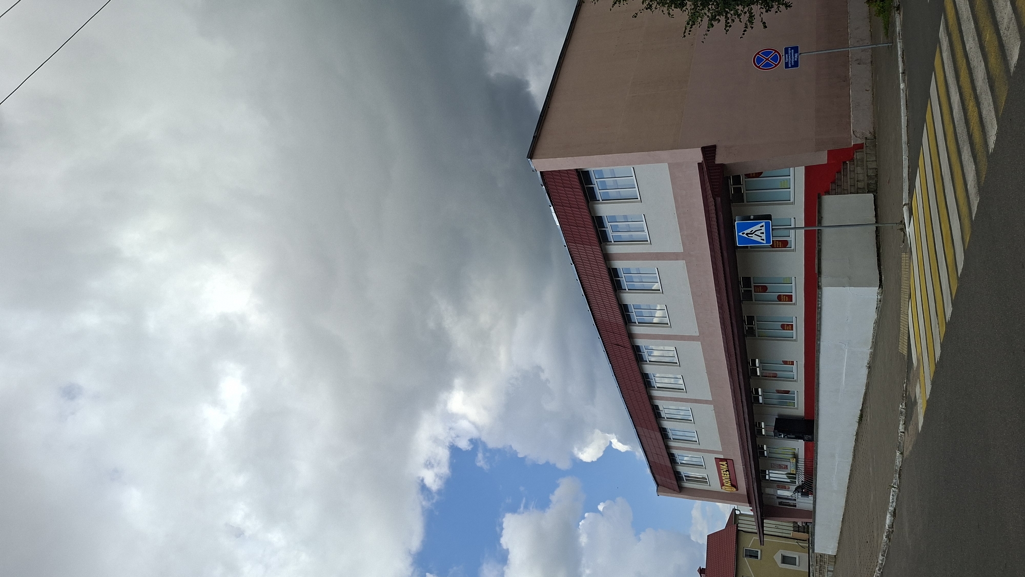 The image depicts a two-story building with a red tiled roof and pale pink and white facade. The building's lower level has a dark red trim below a row of windows. Above this, on the second floor, there is another row of windows. A sign with yellow lettering that reads "КОПЕЕЧКА" is visible on the front of the building. A pedestrian crossing with yellow and white stripes is in the foreground. To the right of the building, there are two blue traffic signs; one depicts a pedestrian symbol, and the other is circular with a red border and a diagonal red line, indicating a prohibition, with text below. The sky is partly cloudy with dramatic grey storm clouds dominating the upper portion of the frame and patches of blue sky visible on the left. A portion of another building with a red roof is visible to the left of the main building. The scene appears to be on a street in the city of Myadzyel, Belarus.