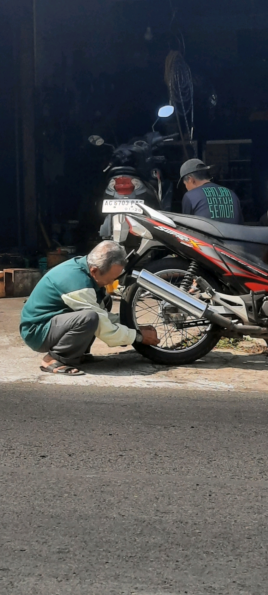 In Selopuro, Indonesia, an elderly man in a green and white jacket and grey pants is crouched down, tending to the rear tire of a motorcycle. He is wearing black and brown sandals. The motorcycle, a red and black "Supra X" model, has a license plate that reads "AC 6703 PS" and "01-26". In the background, another person wearing a dark t-shirt with green text that reads "BANJAR UNTUK SEMUA" and a grey baseball cap is seated on another motorcycle. The scene appears to be outdoors, possibly at a repair shop or roadside stall, given the presence of the motorcycles and the man's action. The ground is paved, and the background is a dark, slightly out-of-focus wall or building. The lighting suggests it is daytime, with strong shadows cast by the sun.