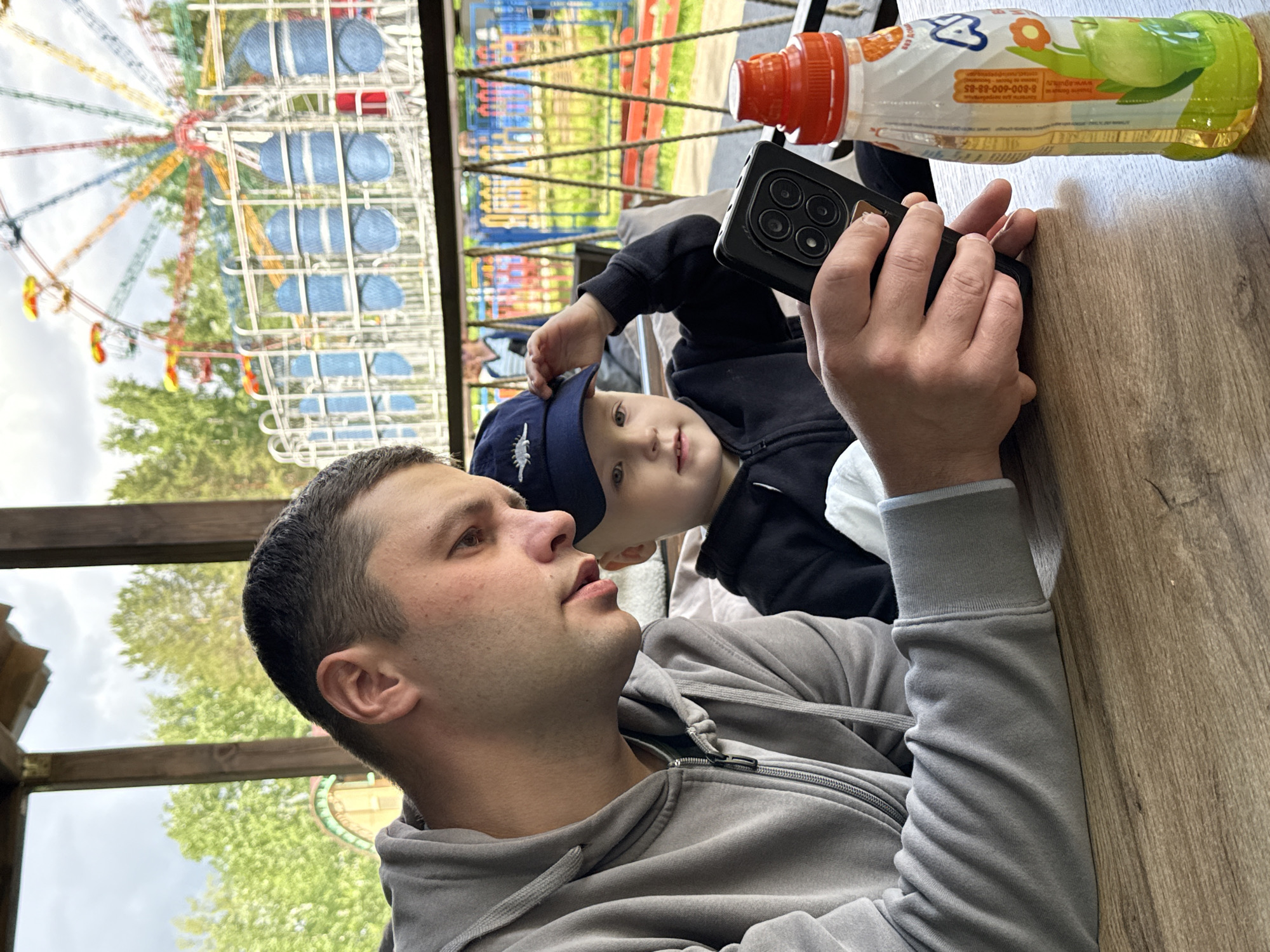 A man and a young child are at a table, inside a covered structure, possibly a gazebo, at what appears to be a fair or amusement park in Chaikovsky, Russia. The location can be inferred from the text on the juice bottle which contains the Russian phone number 8-800-600-88-85. A partially visible Ferris wheel can be seen through the framework above the table. The man, wearing a grey zip-up hoodie, is holding a cell phone with multiple cameras in his right hand. The child, wearing a navy blue baseball cap with a white emblem, is beside him, partially obscured by the phone. An open bottle of juice is also on the table. The table surface is wooden.
