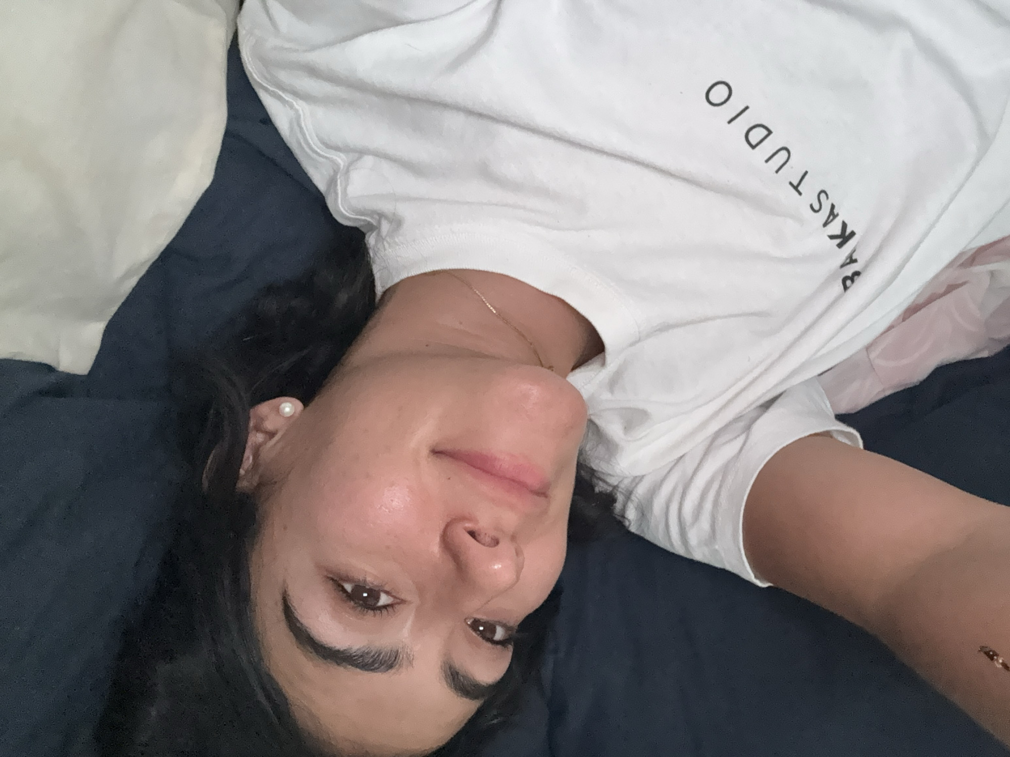A woman lies on a dark blue bed. She is wearing a white t-shirt with the words "RAKASTUDIO" printed on it. She has dark hair, brown eyes and a pearl earring on her left ear. She is wearing a gold necklace.
There is a white pillow to the left of her head.
This scene does not appear to be in Puebla, Mexico.
