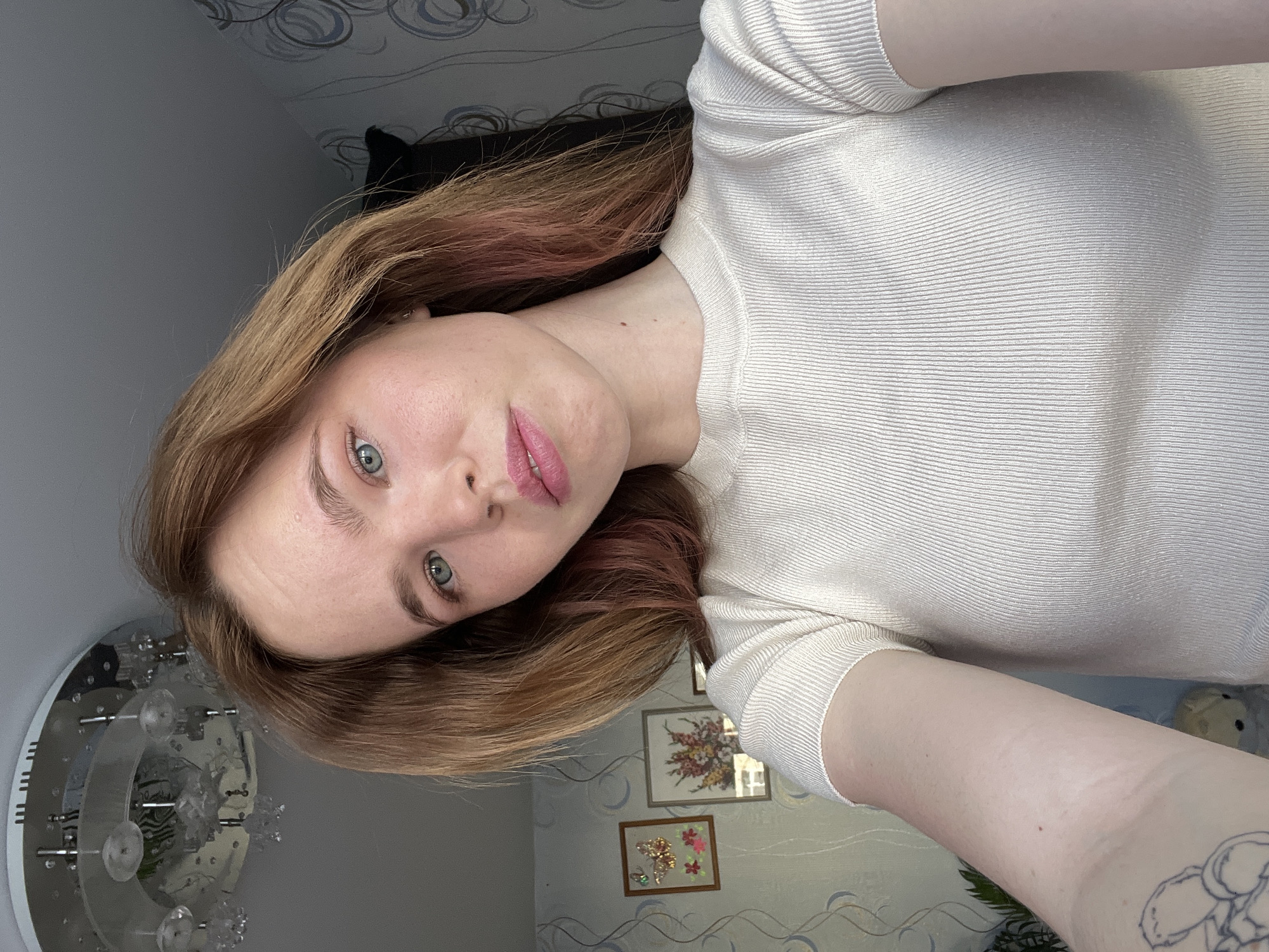 A selfie taken by a fair-skinned woman with light brown hair with some pink highlights. She is wearing a cream-colored ribbed top. The background includes a wall with a light blue patterned wallpaper, framed pictures of floral arrangements, and a modern light fixture. The location is in the city of Chaikovsky, Russia.